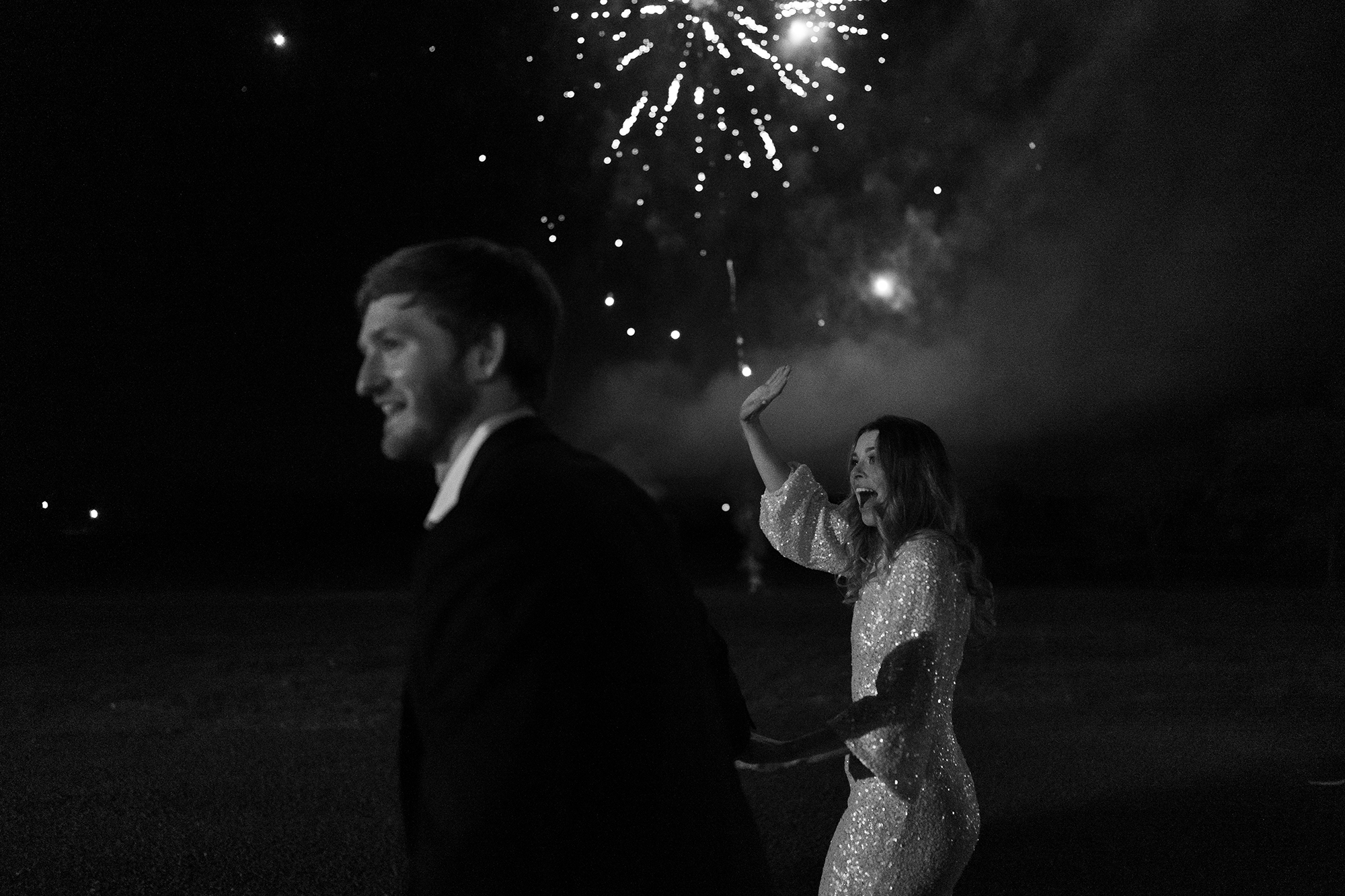 The bride and groom waving to their guests as they leave the reception with fireworks behind them.