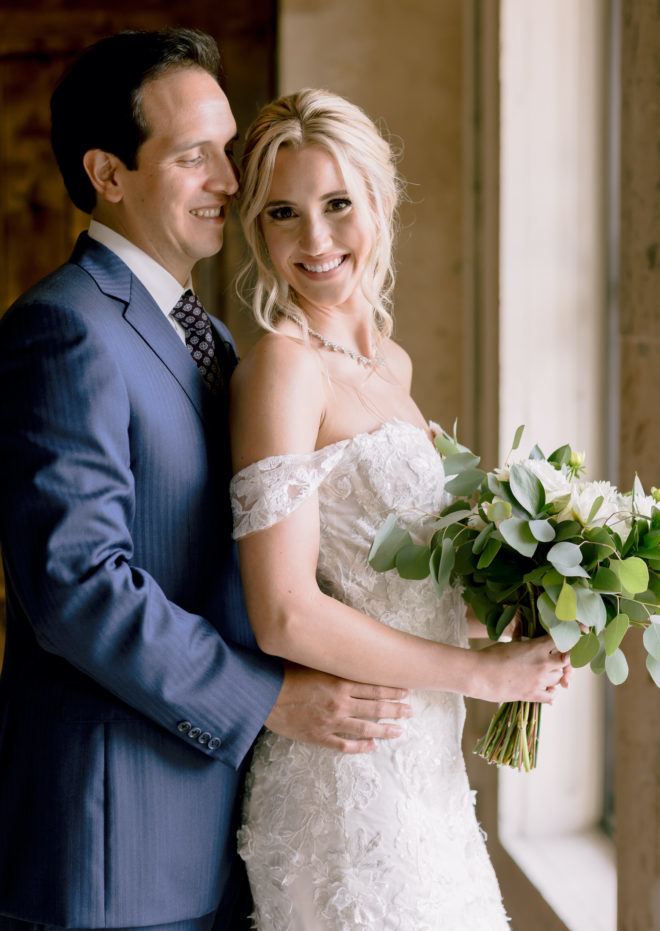 Groom in blue suit and tie holds a blonde bride in an off-the-shoulder gown after their wedding ceremony at wedding venue, The Bell Tower on 34th, in Houston, Texas. 