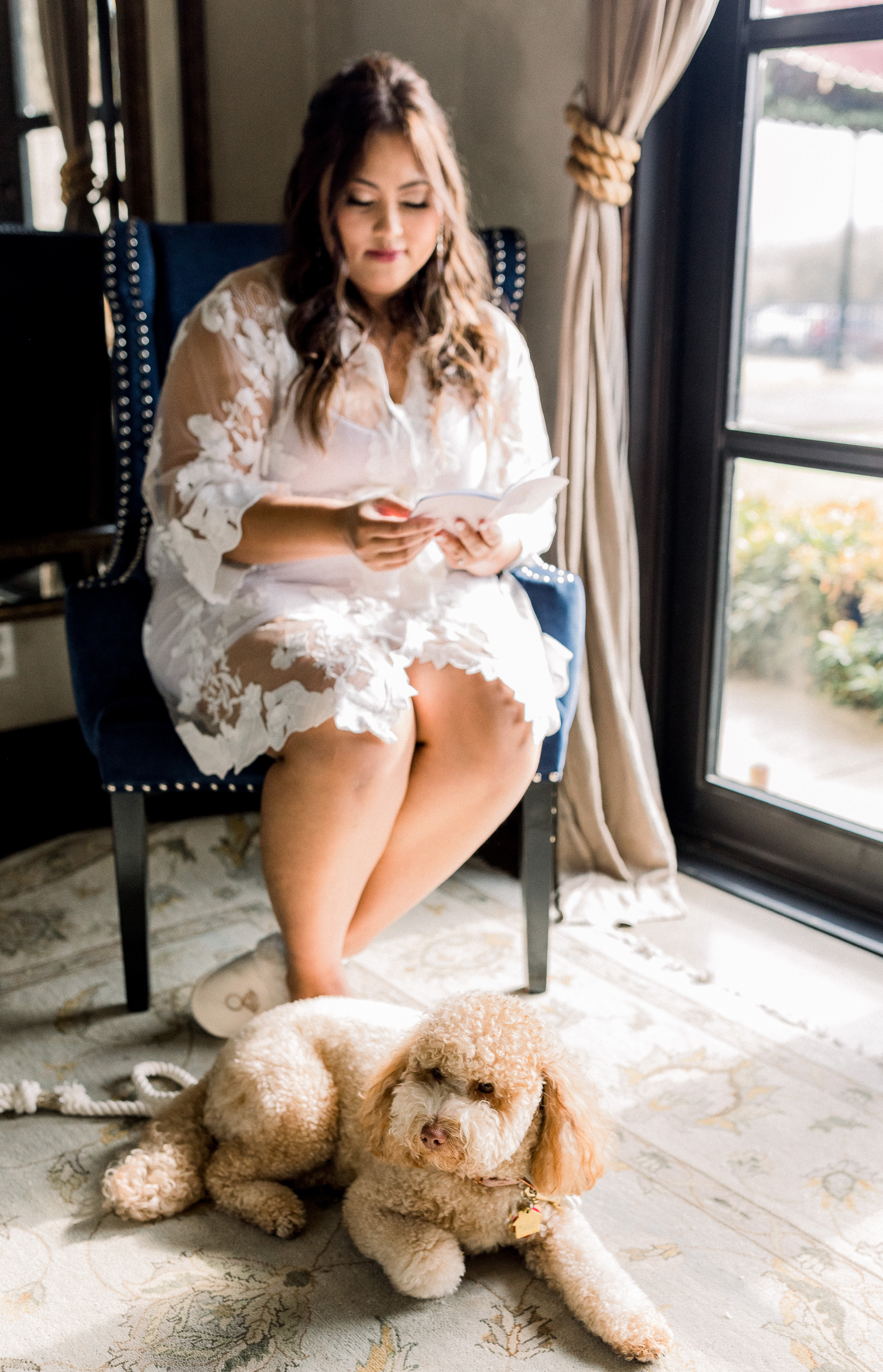 Bride in a lace robe sitting by a window reading her vows with her pet dog at her feet before her wedding ceremony at Ma Maison wedding venue in Dripping Springs, Texas. 