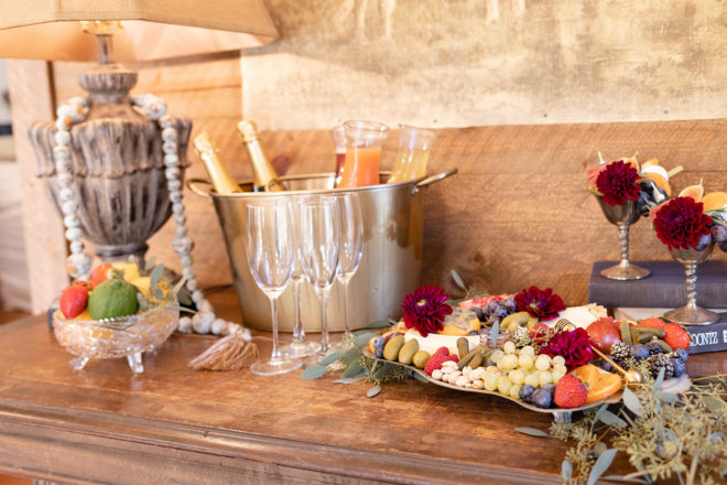 Table top with metal tray with pickles, grapes, olives, strawberries and crimson florals next to a ice bucket of champagne and juice in a guest suite at a Boerne wedding venue and inn. 