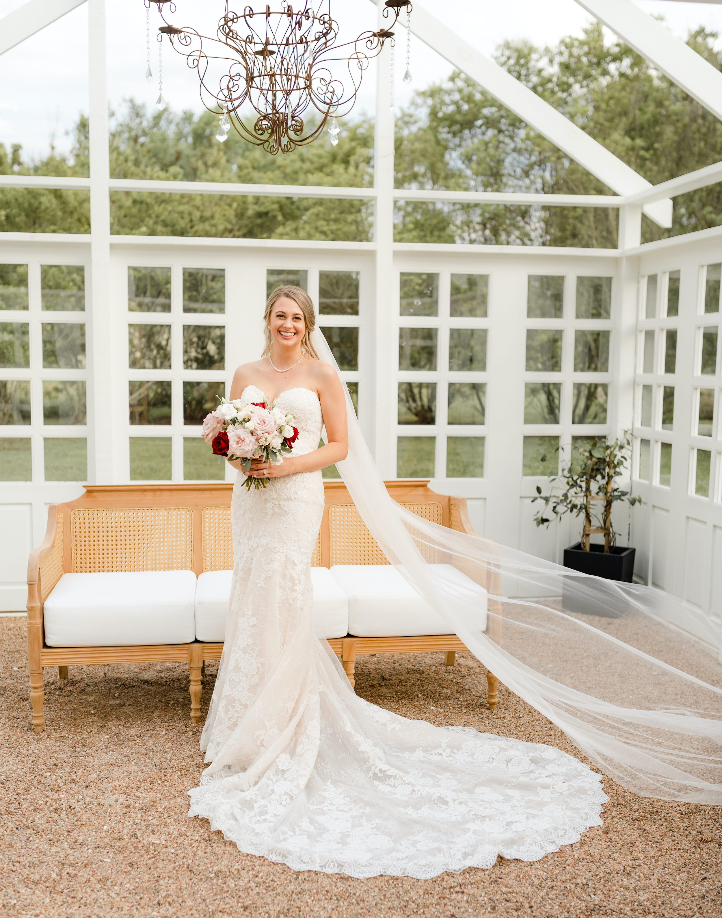 The bride smiles, holding her bridal bouquet and standing inside a greenhouse in her wedding dress.