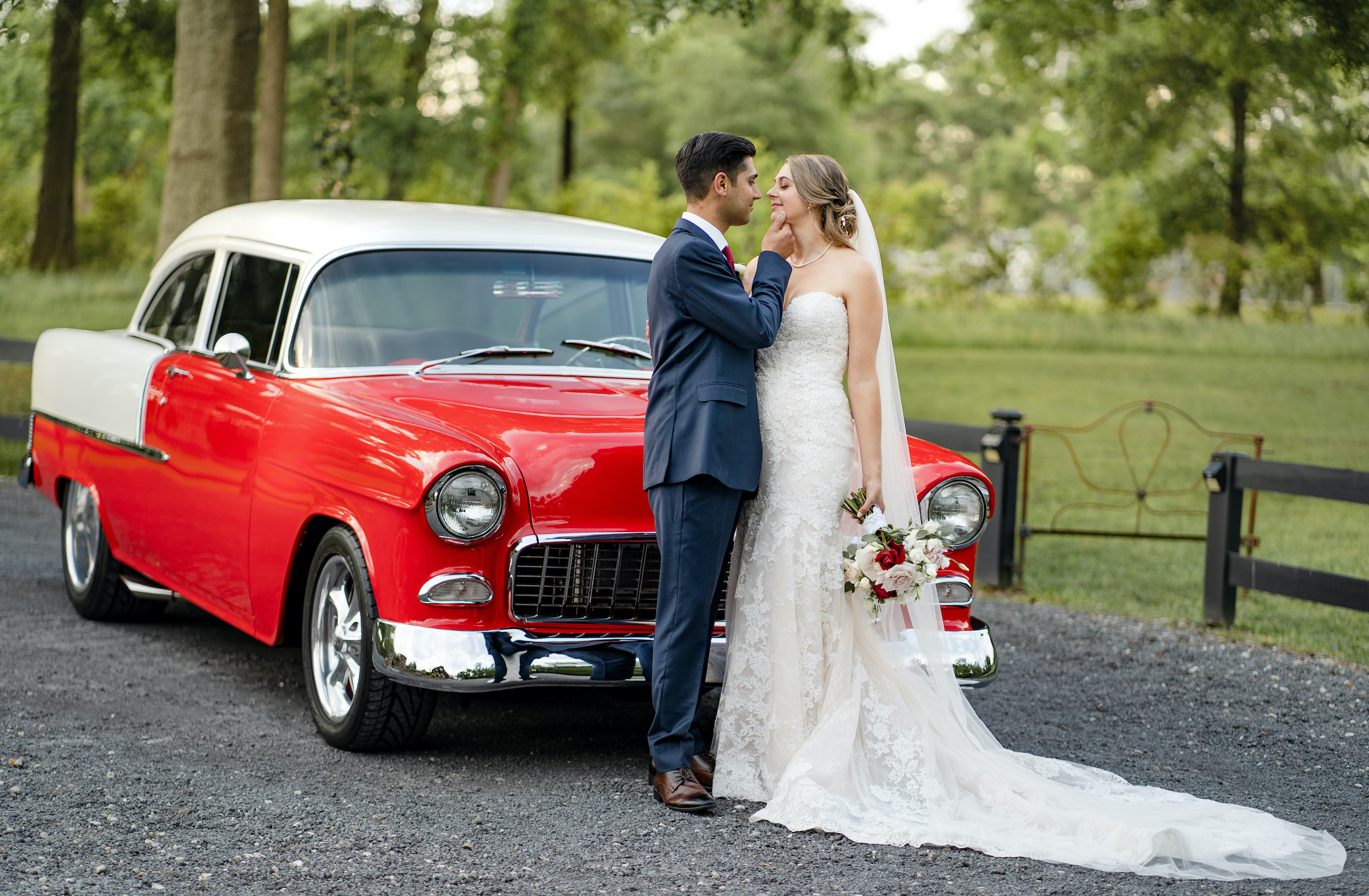 The bride and groom stand in front of a bright red classic car.