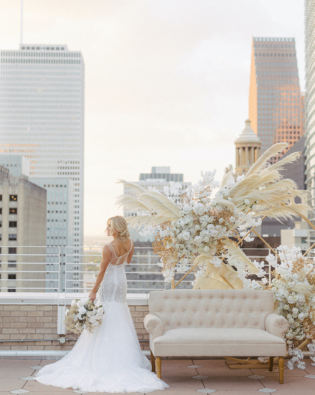 The bride has her back to the camera and holds her bridal bouquet next to the couch, which is decorated in white, gold and beige florals.