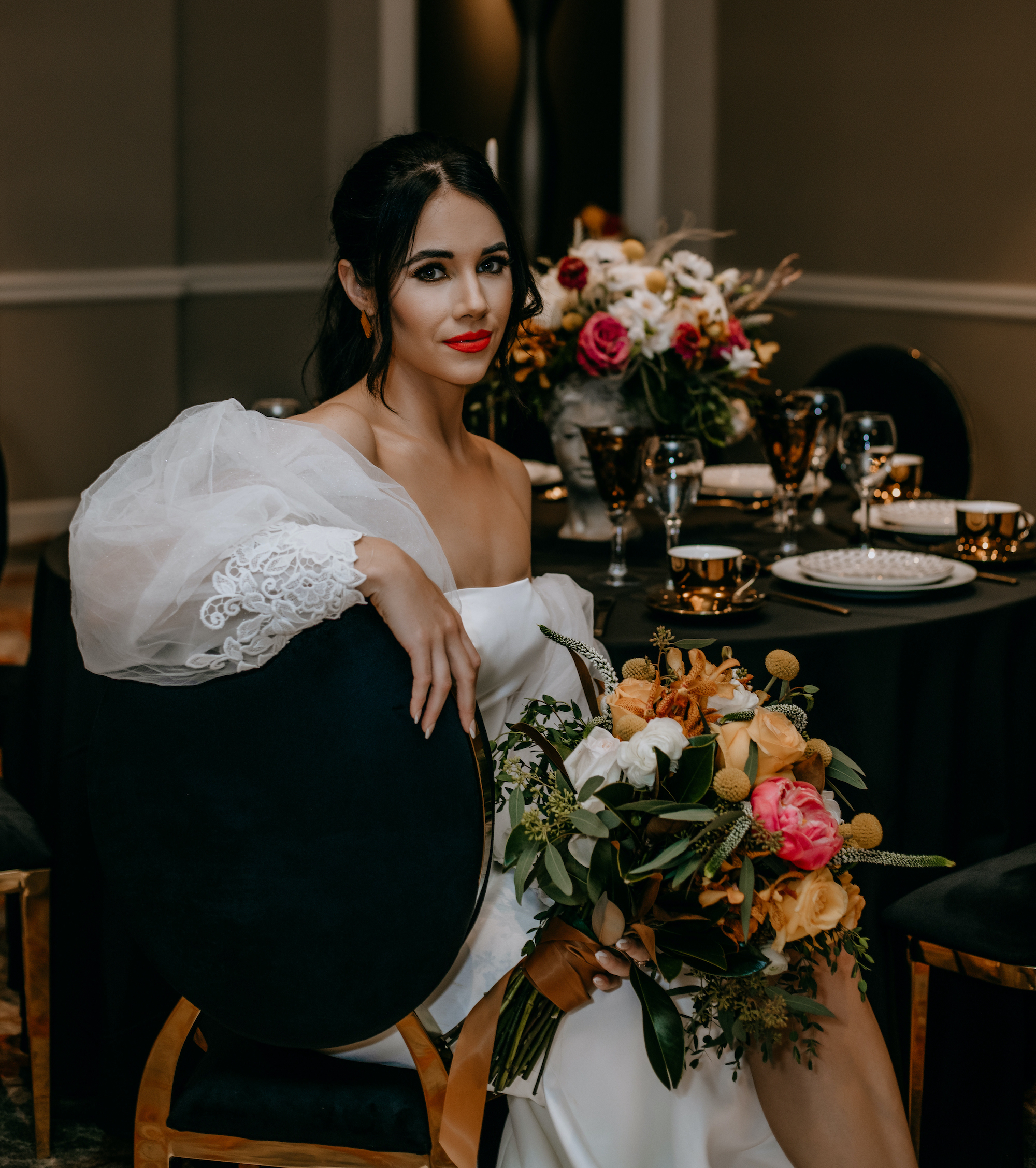 A bride sits at a dinner table set with gold dinnerware and a vibrant flower centerpiece.