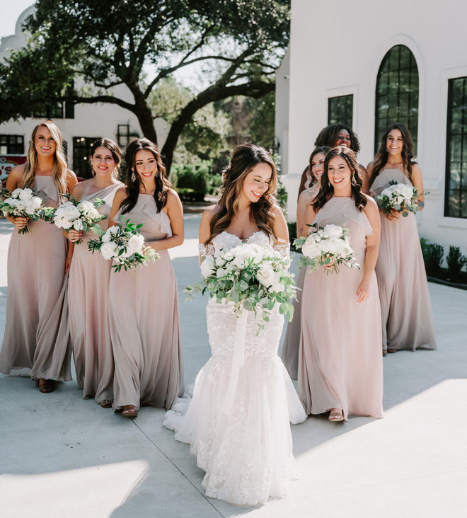 Bridesmaids follow the bride outside of the wedding venue. They are all holding their flower bouquets full of greenery and ivory blooms.