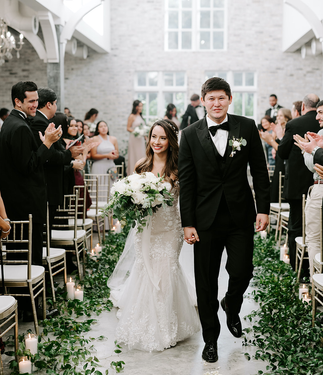 The bride and groom happily walk down the aisle surrounded by their guests clapping. Lush greenery trails down the aisle on either side of them.