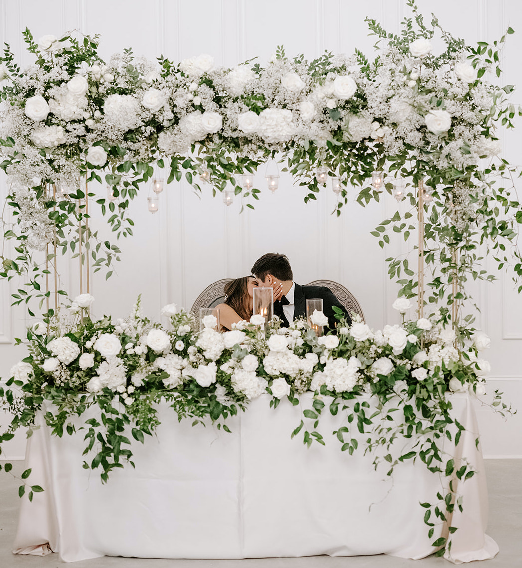 The bride and groom kiss at their table at the reception. Lush greenery and ivory flowers adorn the table for the whimsical wedding.