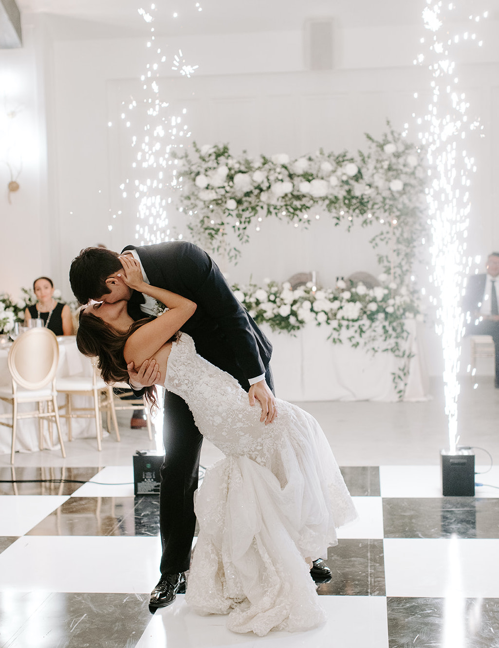 Groom leans bride over for a kiss on a black and white checkered dance floor with sparklers behind them at their wedding reception at The Peach Orchard Venue in the Woodlands, TX