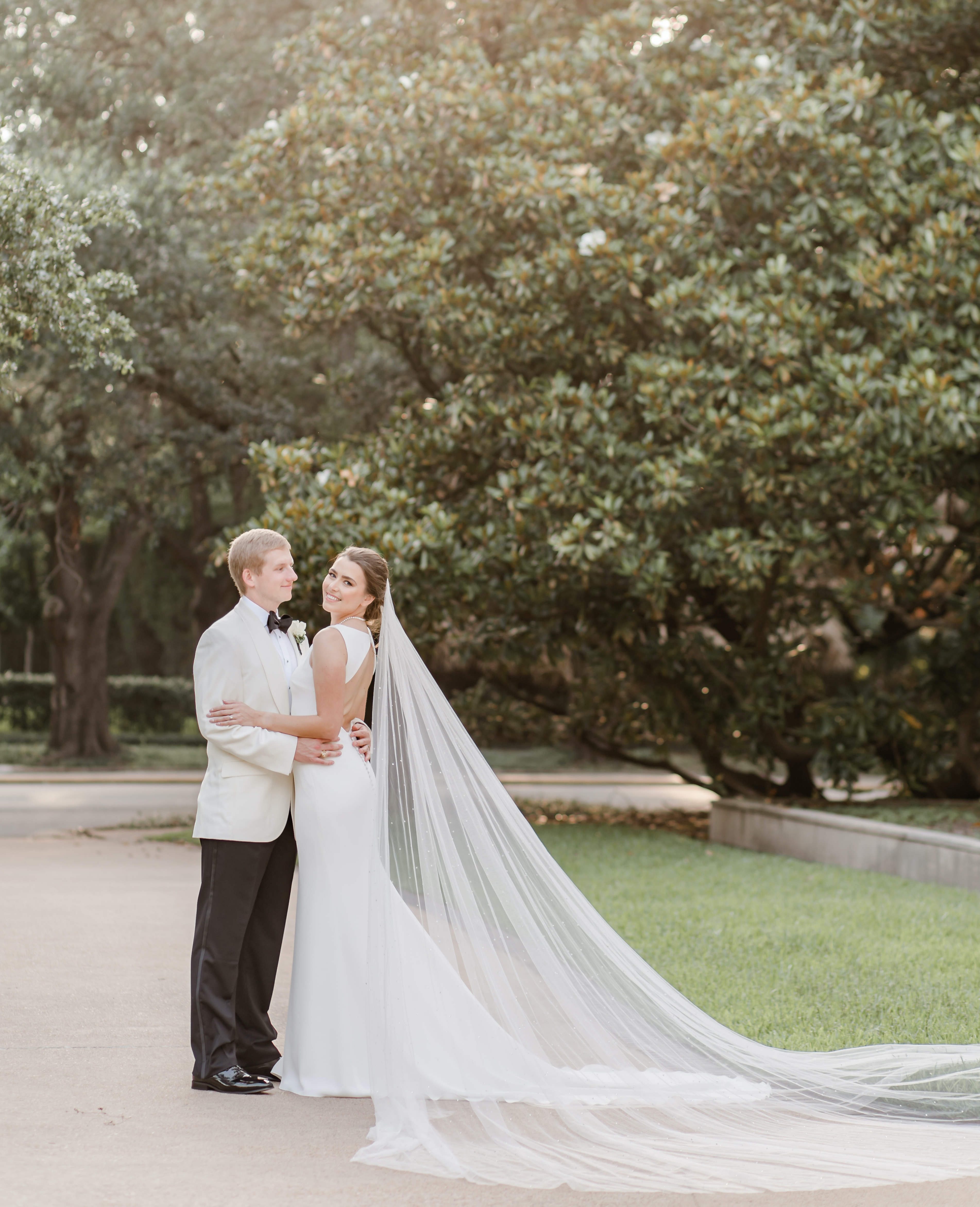 The groom looks at his bride as she looks at the camera. Her veil cascades to the ground outside in the Houston museum district.