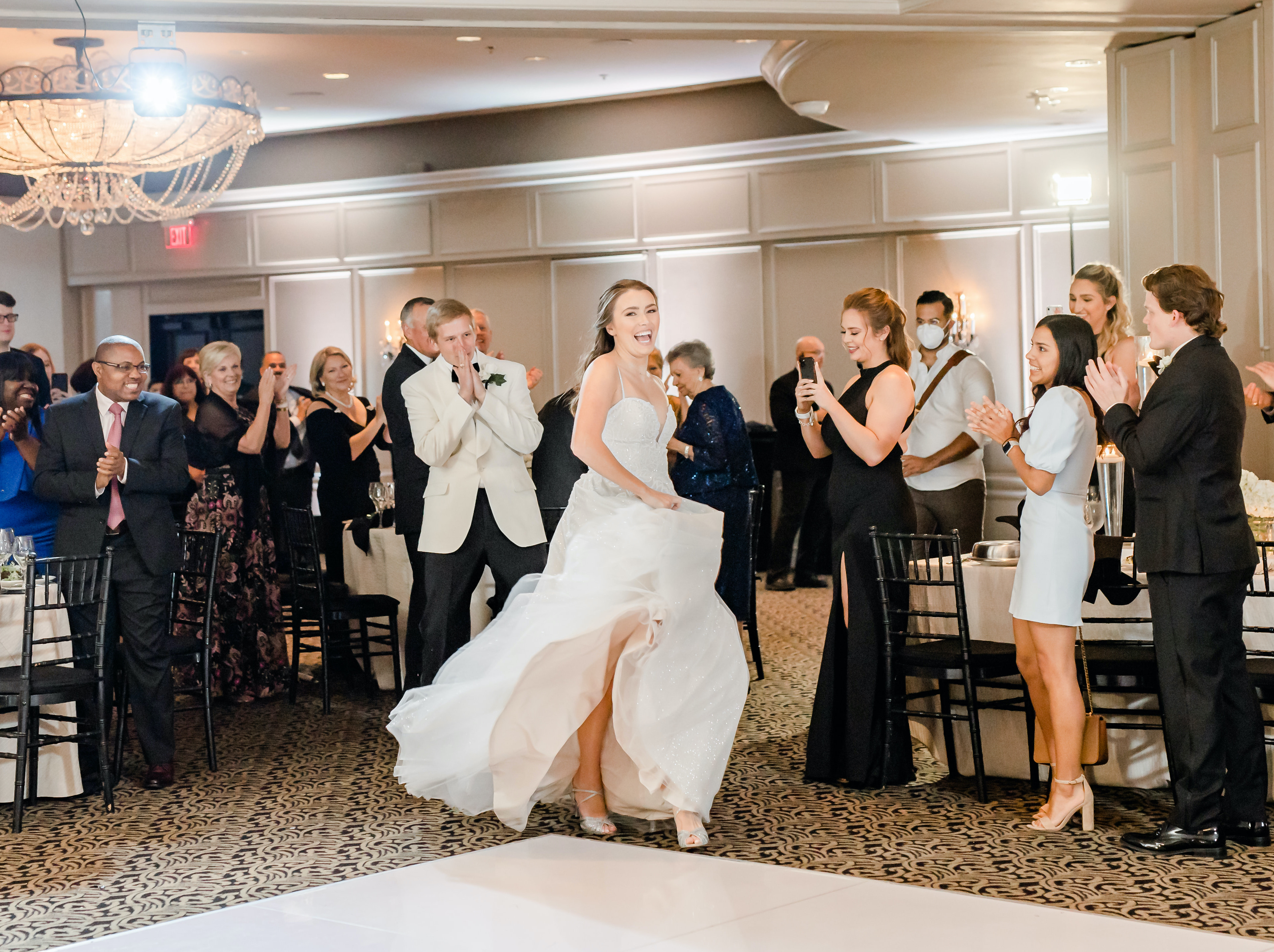 The bride runs to the dance-floor in a sweetheart gown while the guests clap and her husband stands behind her. They are in the Houston museum district for their reception.