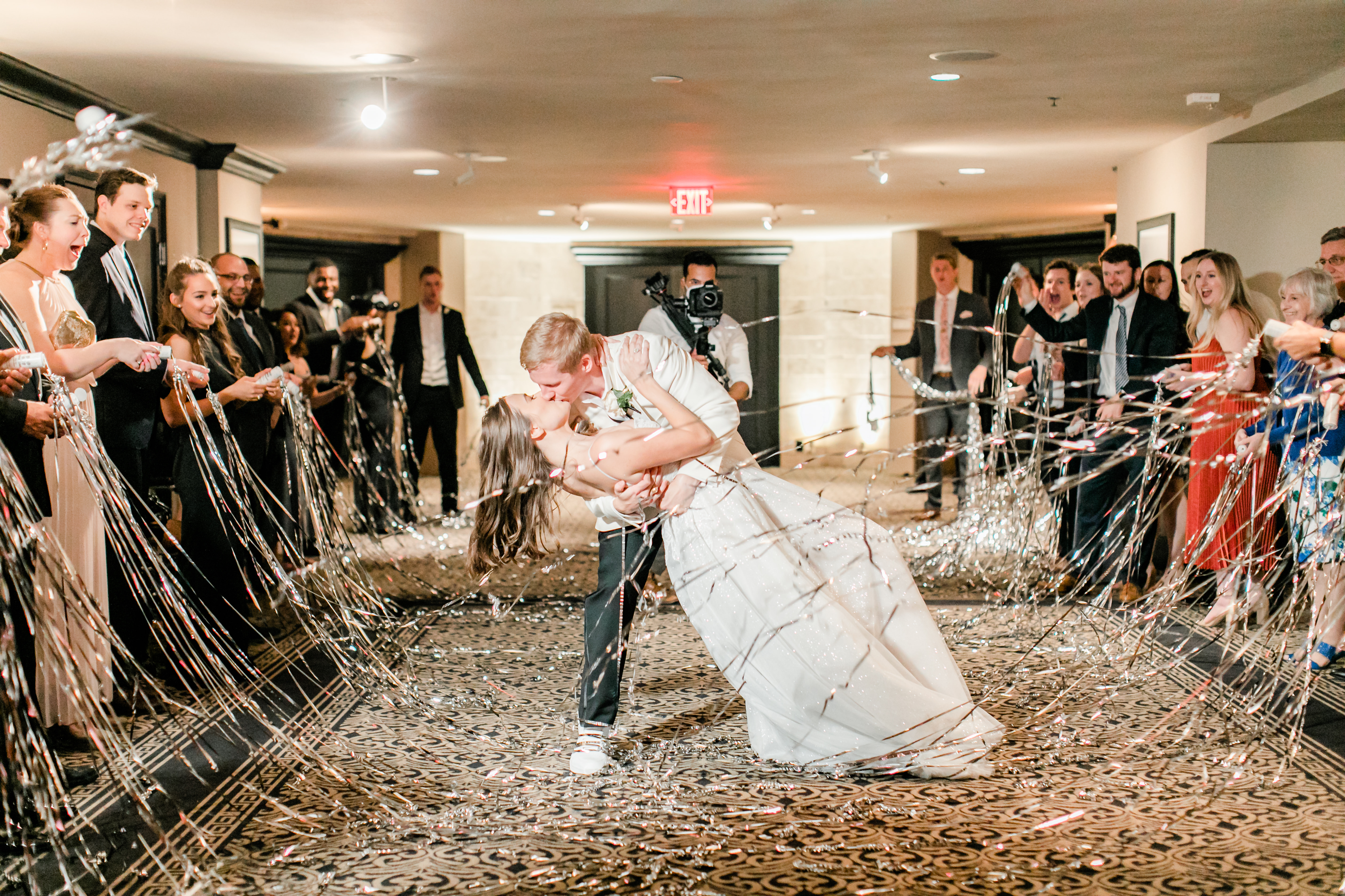 The groom dips his bride and they kiss at their wedding send-off in the Houston museum district while their guests pop silver streamers and cheer them on.