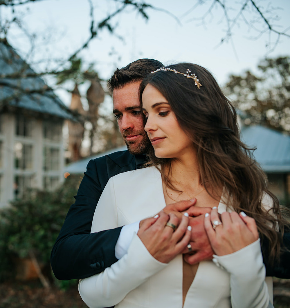 The groom hugs his bride as they both look away from the camera. They are standing outside at their winter wedding at 7F Lodge in College Station, TX.