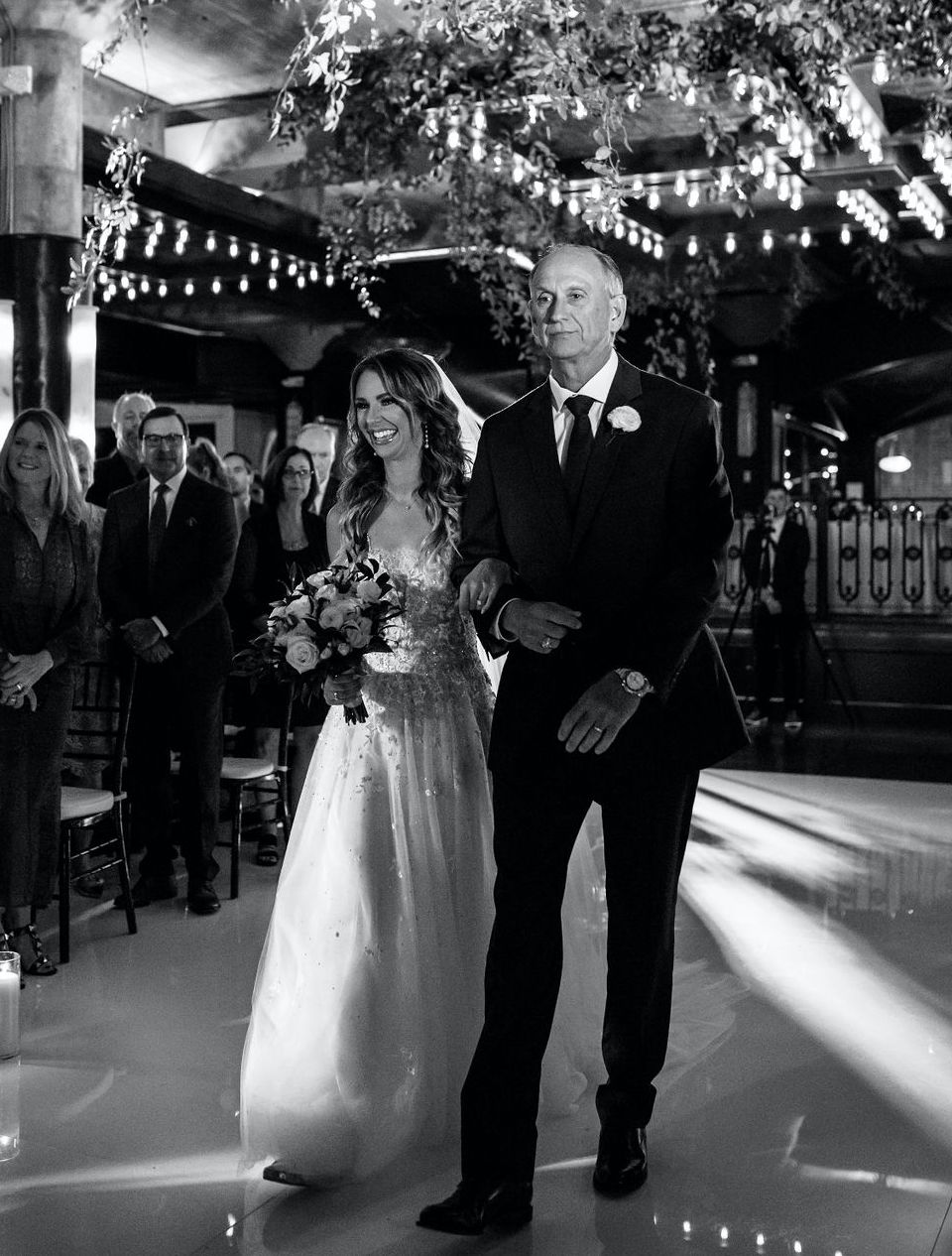 A black and white photo of the bride smiling as she walks down the aisle with her dad at her Astorian wedding in Houston, TX.