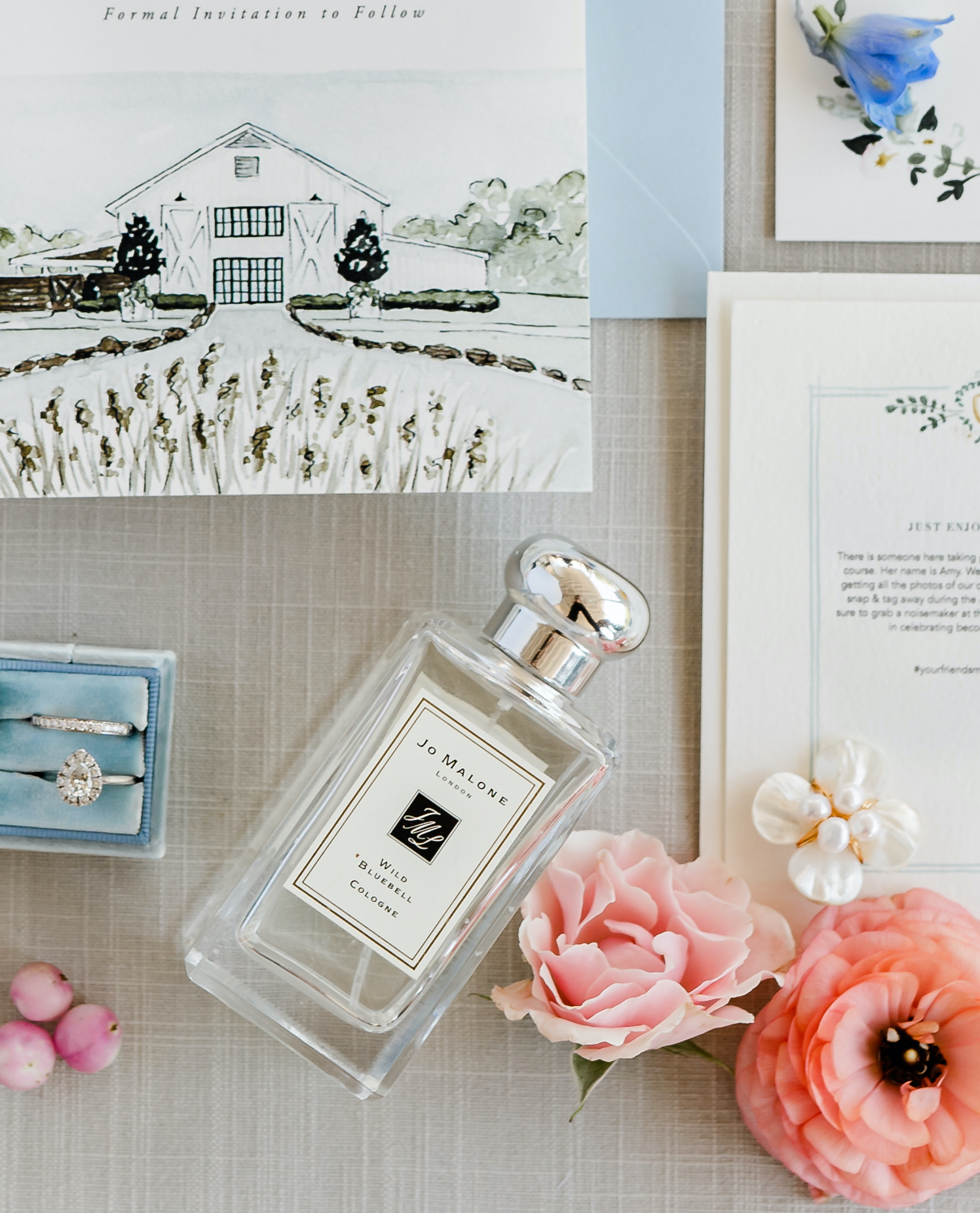 Flat lay of a illustrated wedding invitation suite, a baby blue velvet box with a pear shaped wedding ring and a band, faux pink florals and a bottle of perfume which reads, "Jo Malone London, Wild Bluebell Cologne". 