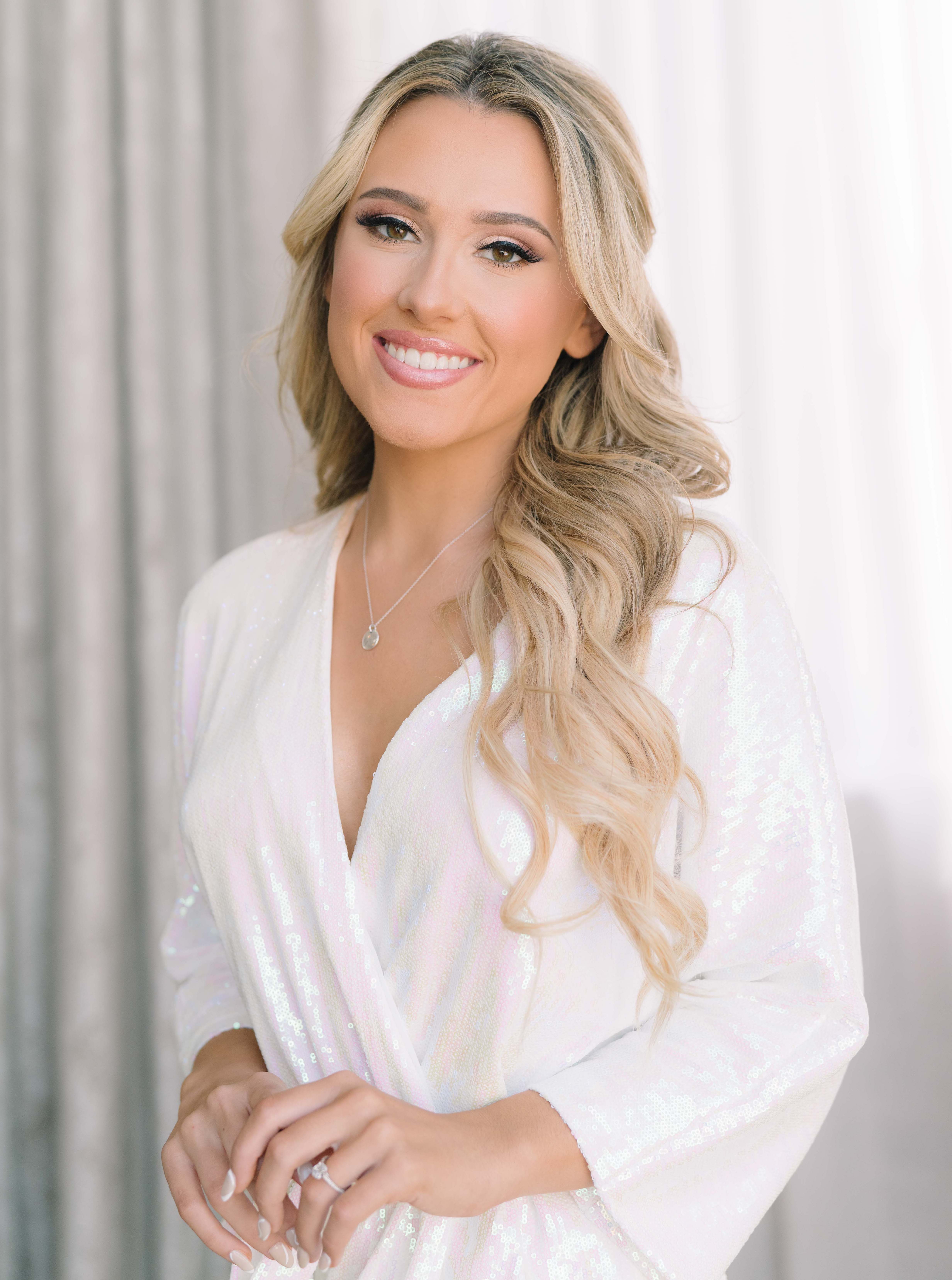 A bride smiles with her hair and makeup done. She is in a white iridescent sequin robe before her Summer Wedding With a Vibrant Palette of Pink.