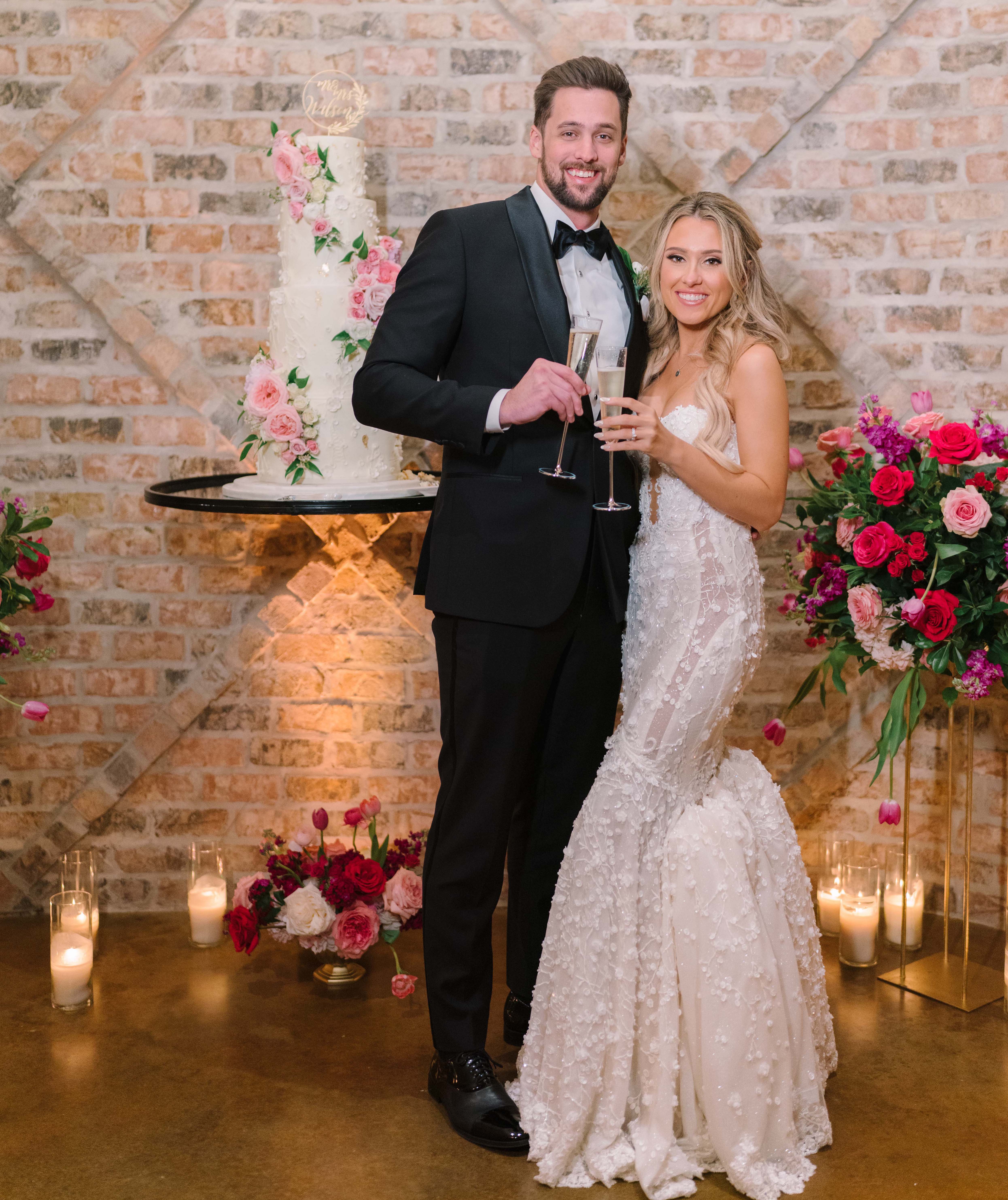 A bride and groom stand with champagne glasses next to their four-tier white wedding cake with pink flowers. Summer Wedding With a Vibrant Palette of Pink