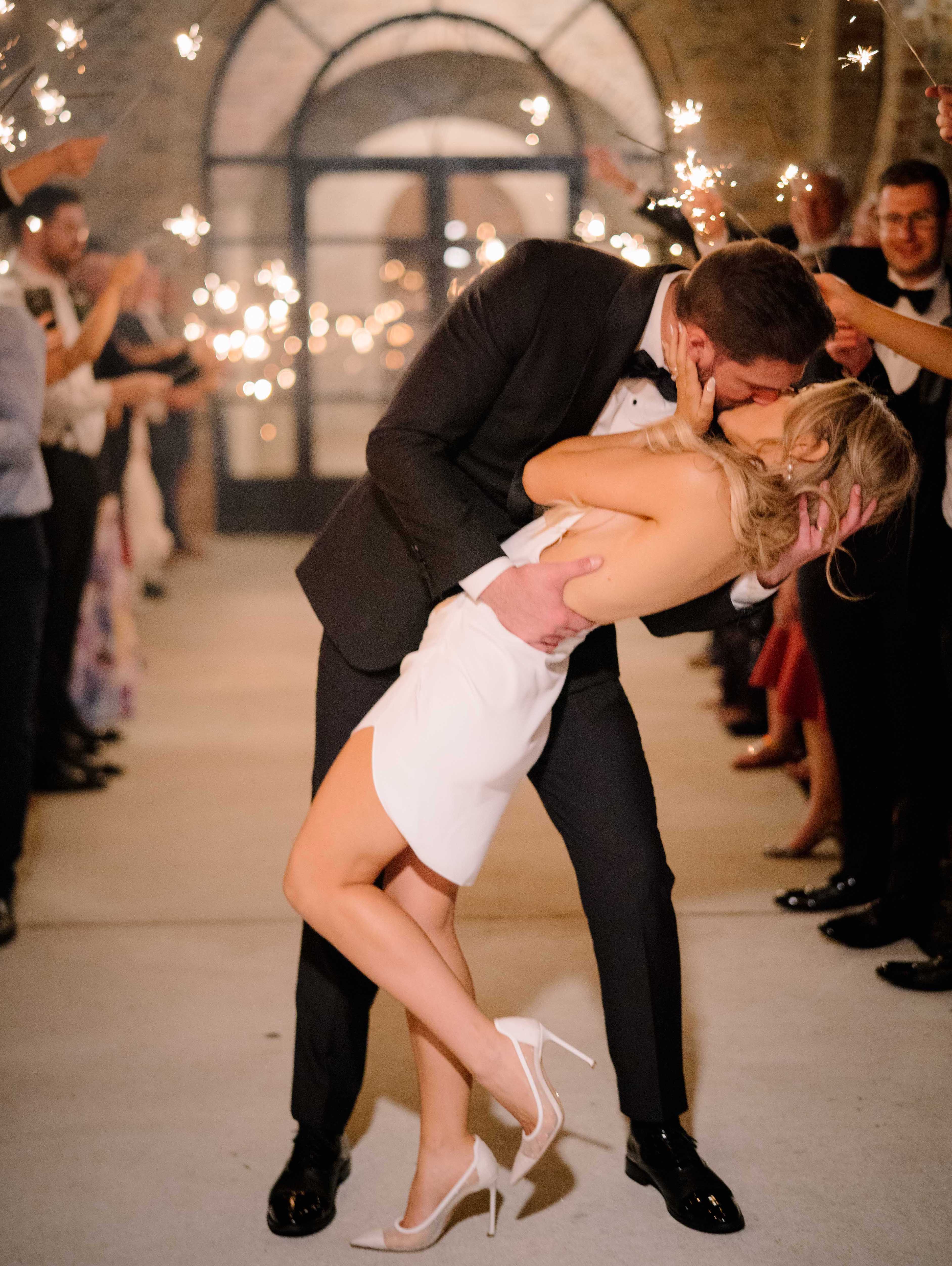 A bride and groom kiss in the middle of an aisle their guests made during their grand exit. Their guests hold sparklers.