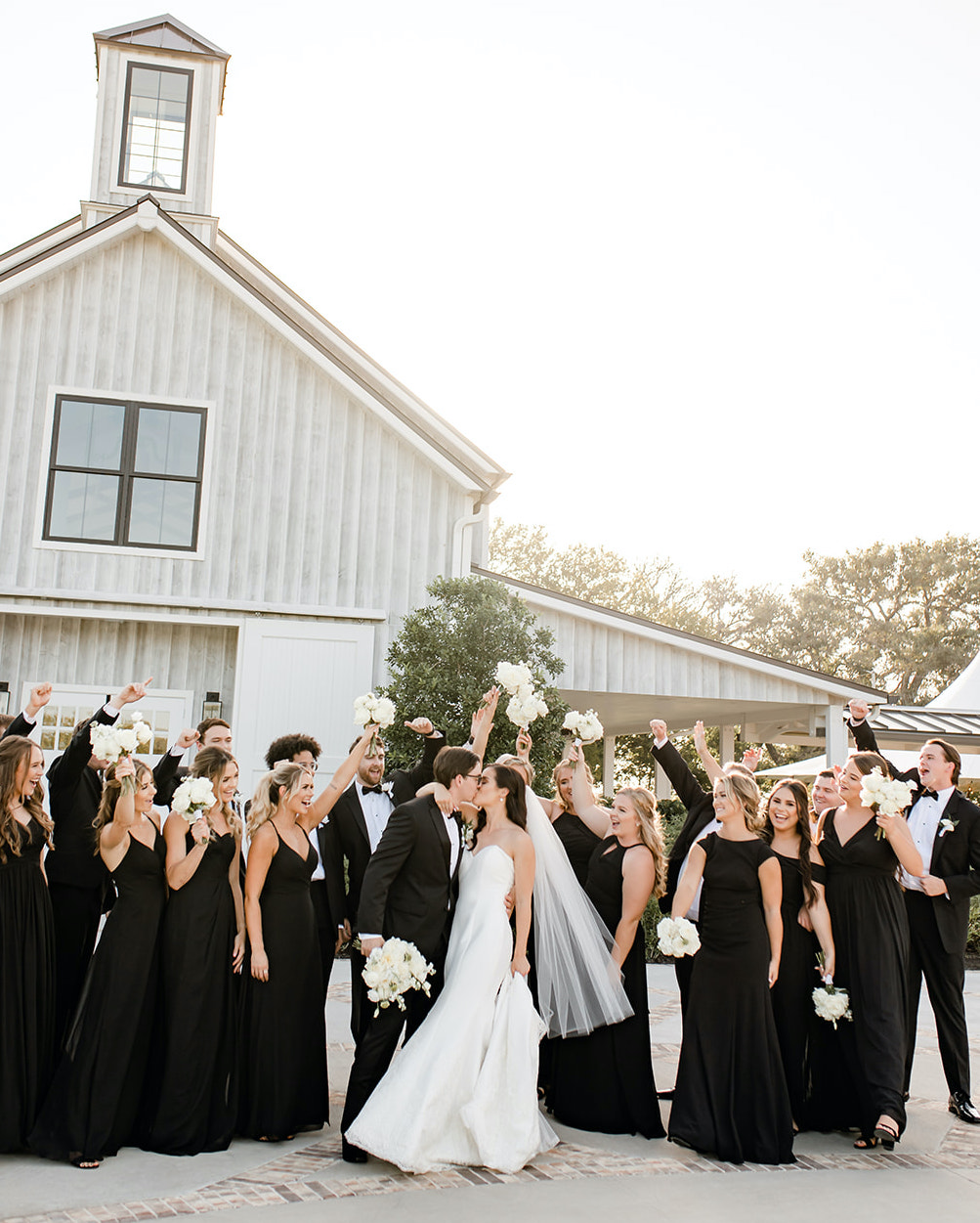 A bride and groom kiss with their wedding party celebrating behind them at their elegant summer wedding in Montgomery, TX.