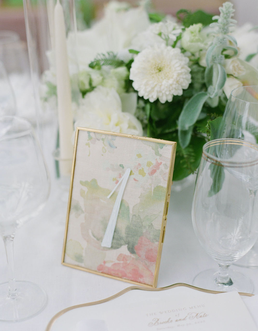 Details on a reception table including a floral centerpiece with greenery and white flowers and a table marker.