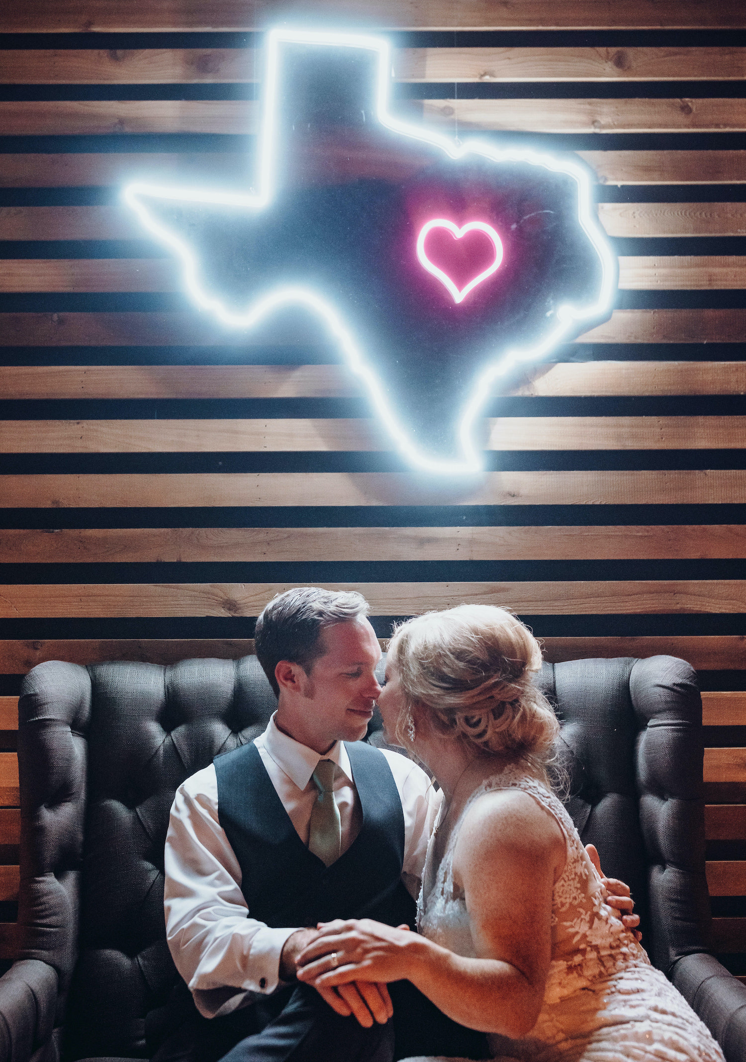 A bride and groom sit in front of a wooden wall with a neon sign shaped as Texas with a heart near Houston.