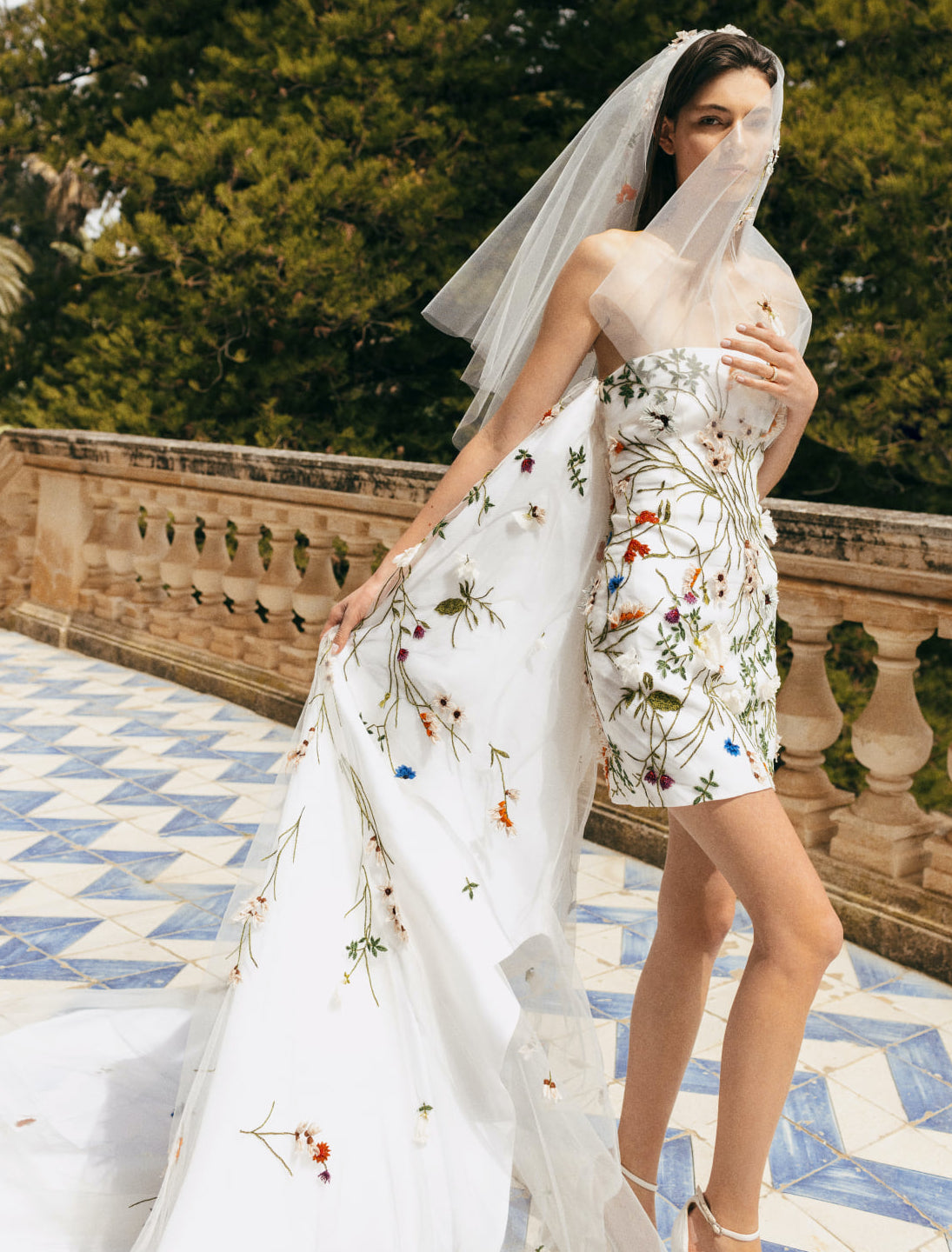 A bride wears a Monique Lhuillier wedding gown that is short and fitted with a flowing train. The strapless wedding dress has colorful embellished flowers covering it.