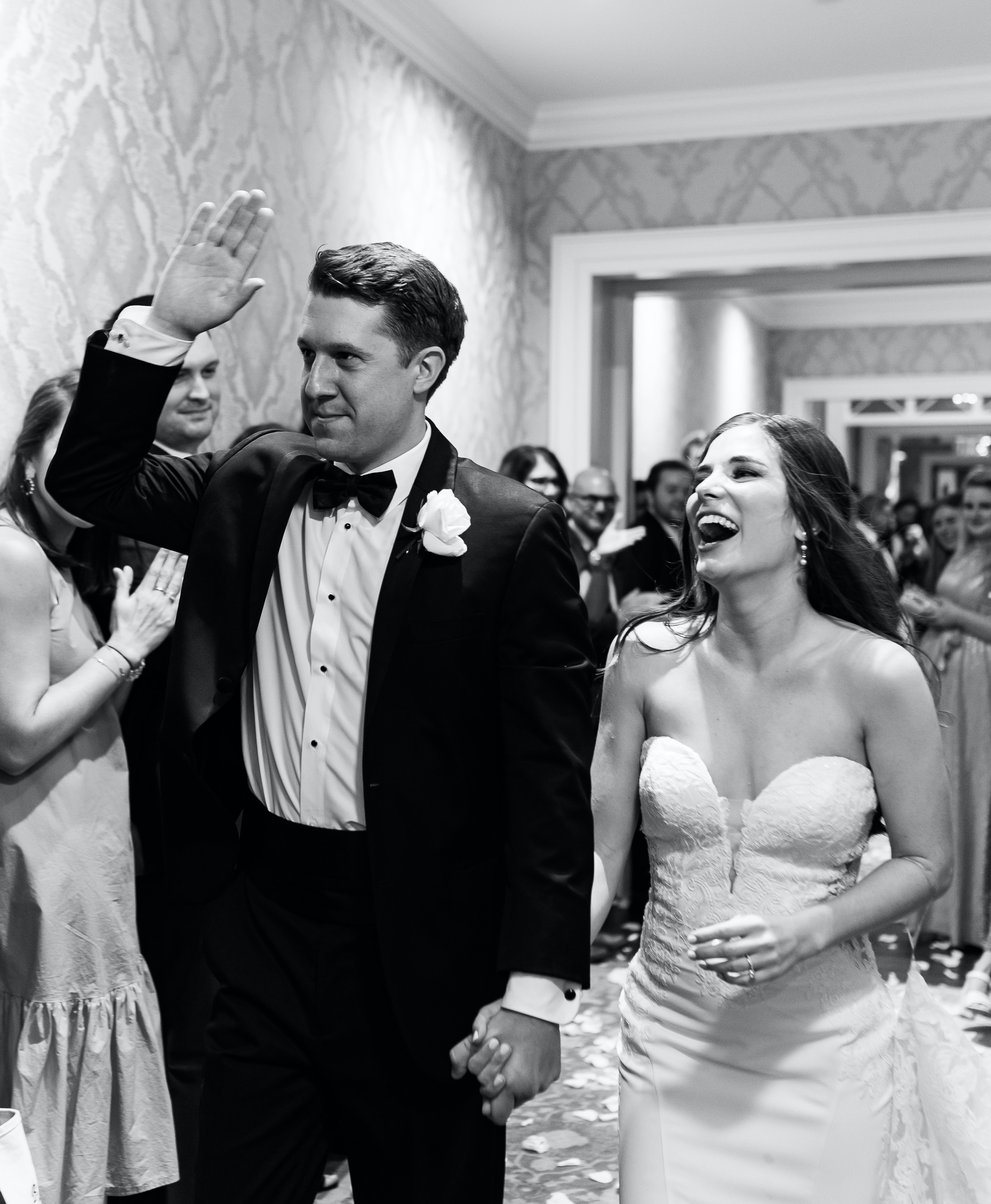 A black and white photo of a bride and groom smiling at their guests and holding hands as they leave their wedding reception.