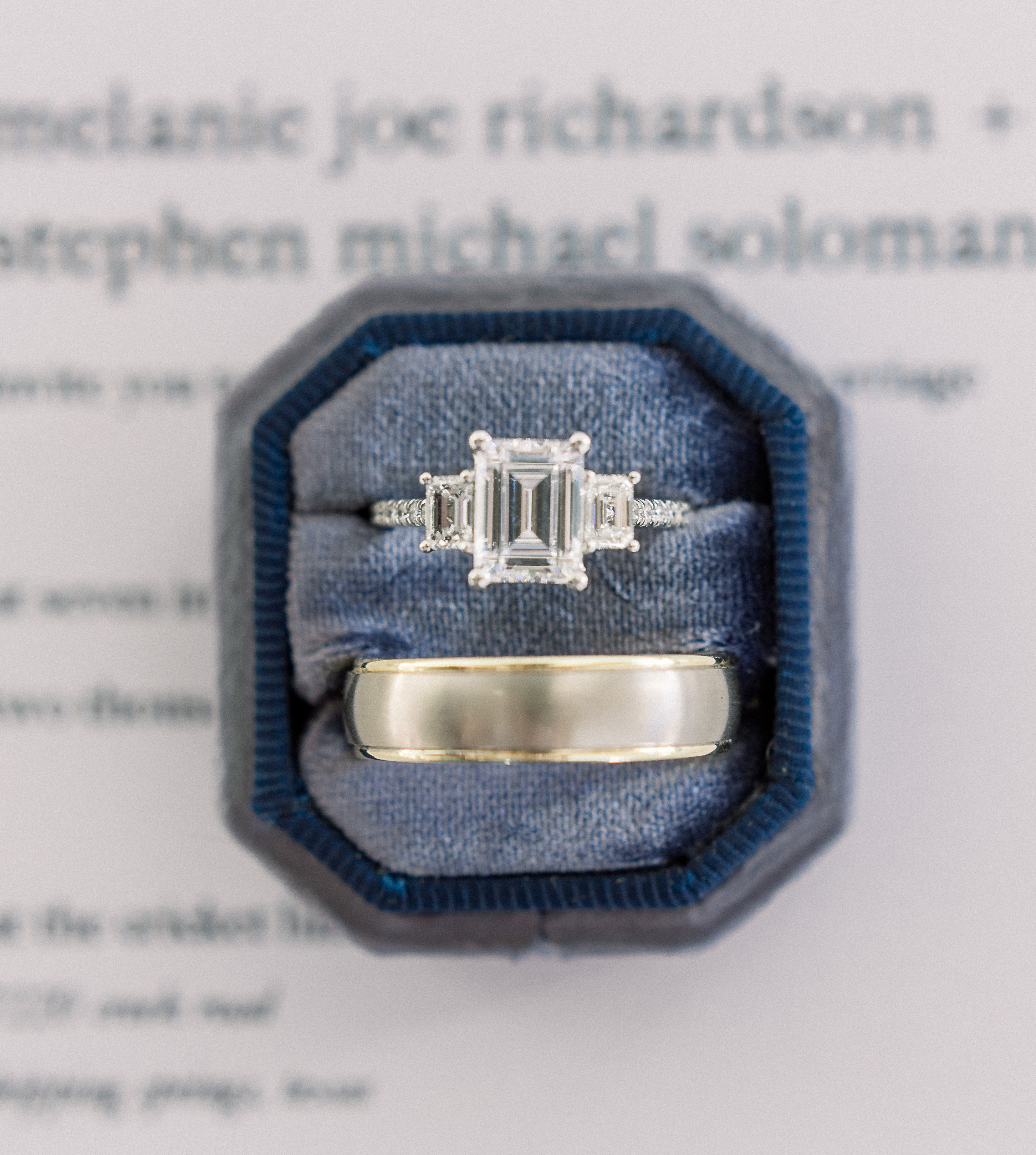 A three-stone engagement ring with an emerald cut is in a navy ring bow next to the groom's wedding band for a hill country styled shoot in Dripping Springs, TX.