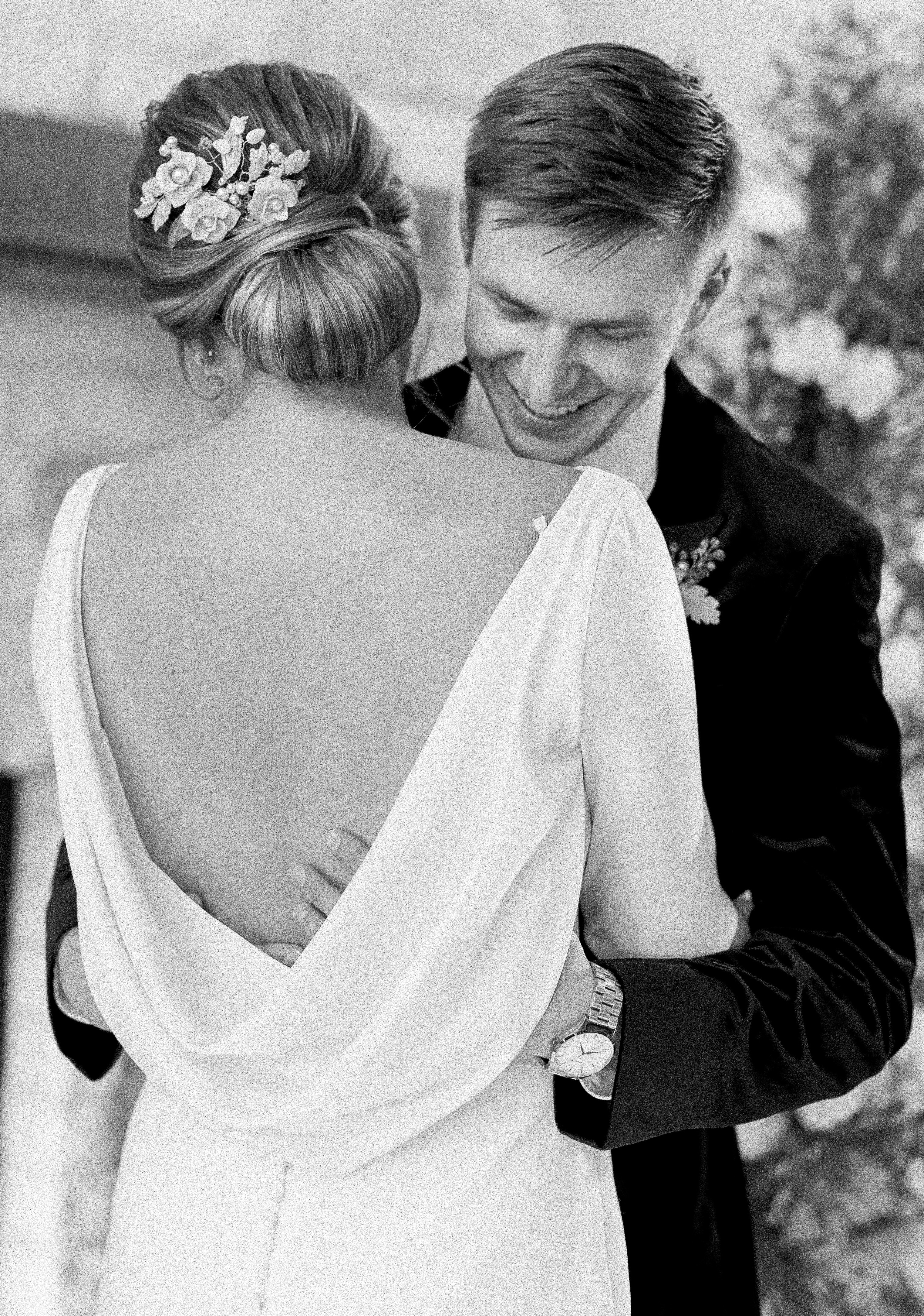 A black and white photo of a groom hugging his bride. Her back is to the camera and she wears a wedding dress with a drooping back.