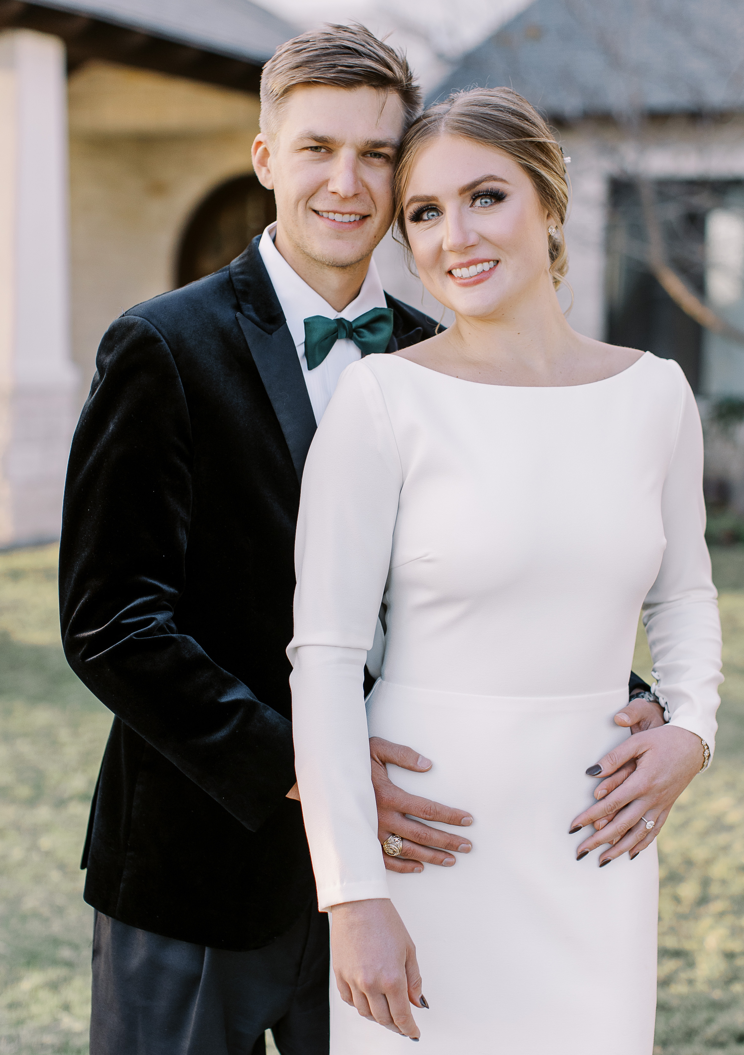 A bride and groom smile at the camera while standing outside the wedding venue, Cricket Hill Ranch in Dripping Springs, TX.