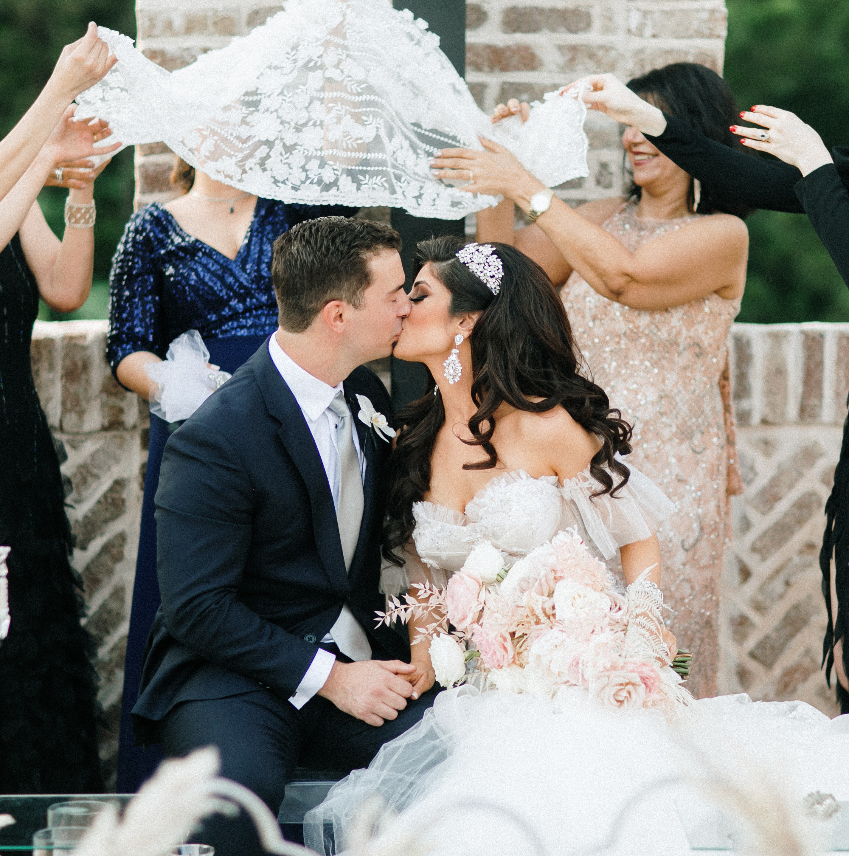 A bride and groom kiss under a blanket held up by their wedding guests at their pink and lilac Persian wedding ceremony.