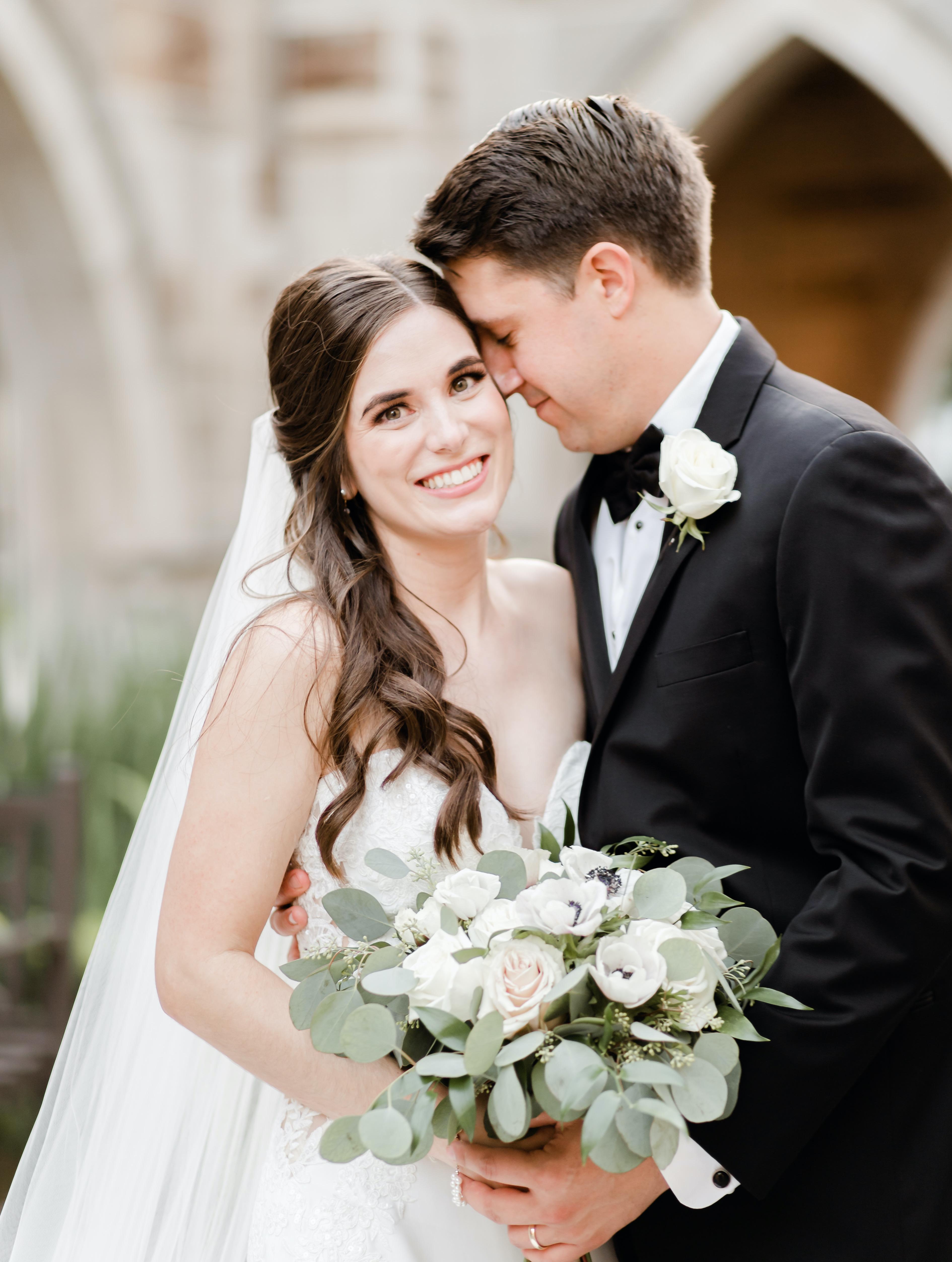 A bride looks at the camera while her groom hugs her on the side and places his forehead on hers.