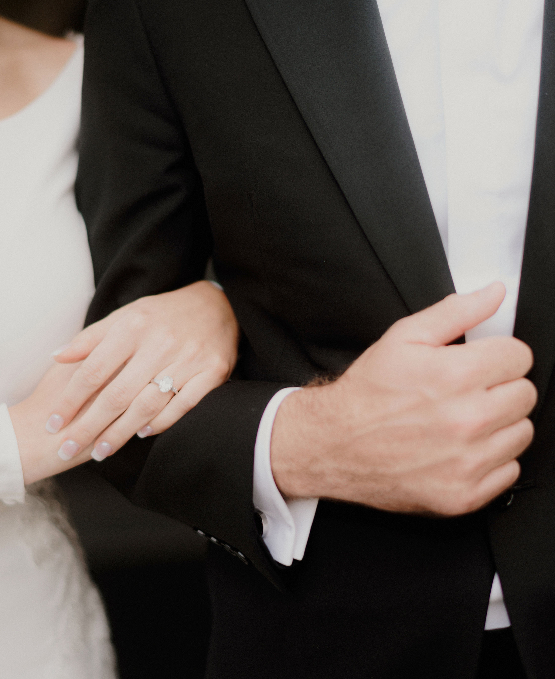 An up-close photo of a bride and groom's hands. The bride wears her oval-cut engagement ring.