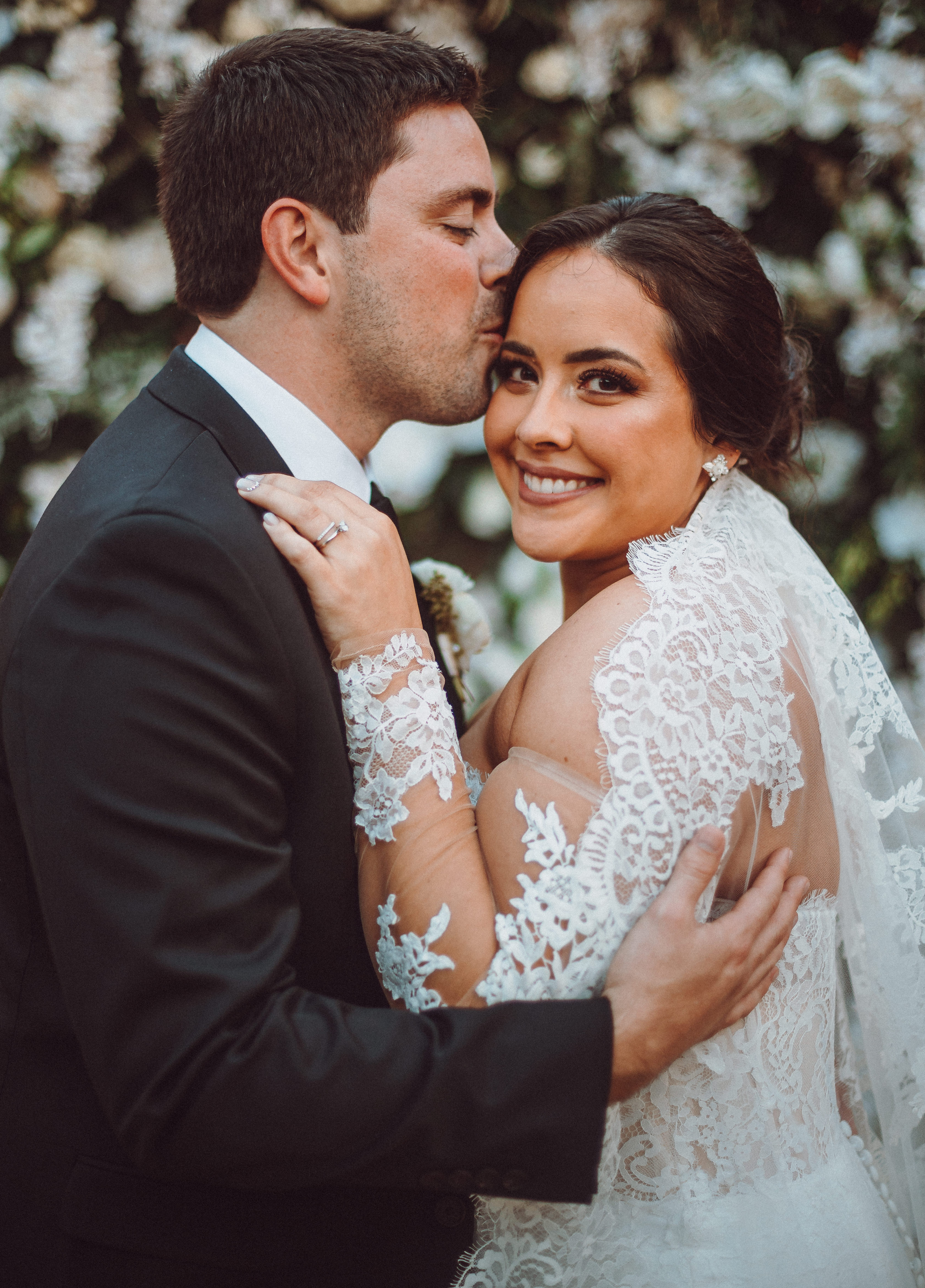 A groom kisses his brides head as she smiles and looks at the camera during their alfresco wedding with terra cotta accents in Houston, TX.