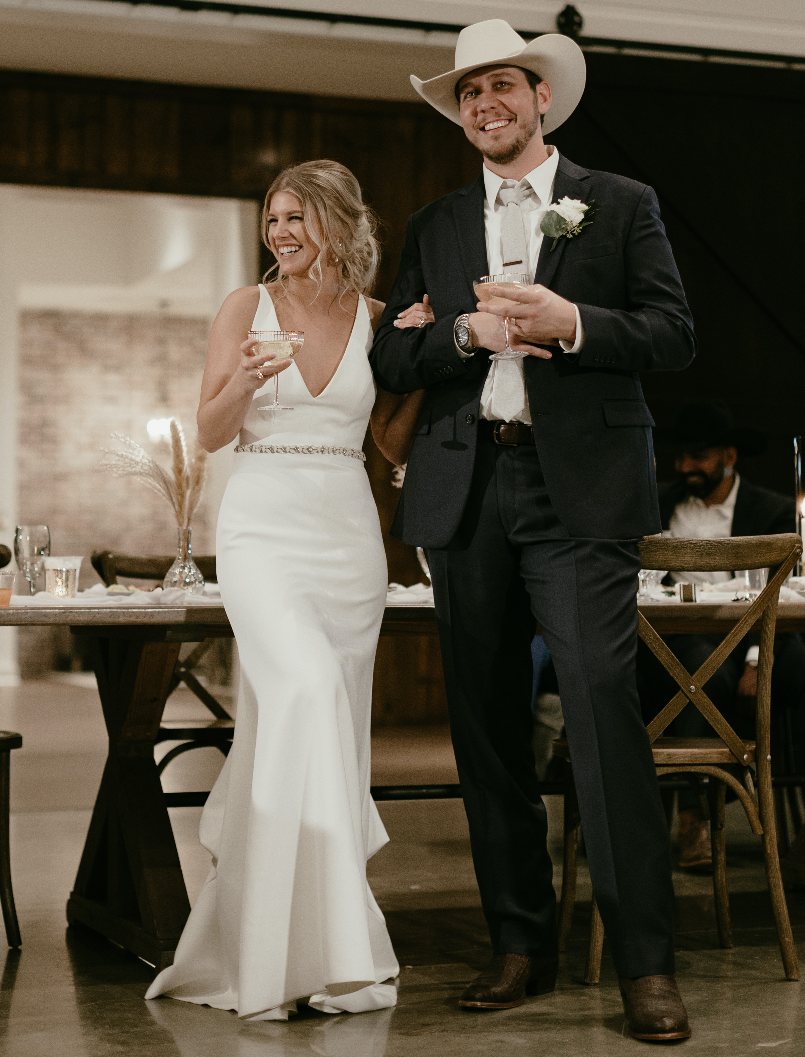 A bride and groom smile while standing and holding champagne glasses at their reception in Brenham, TX.