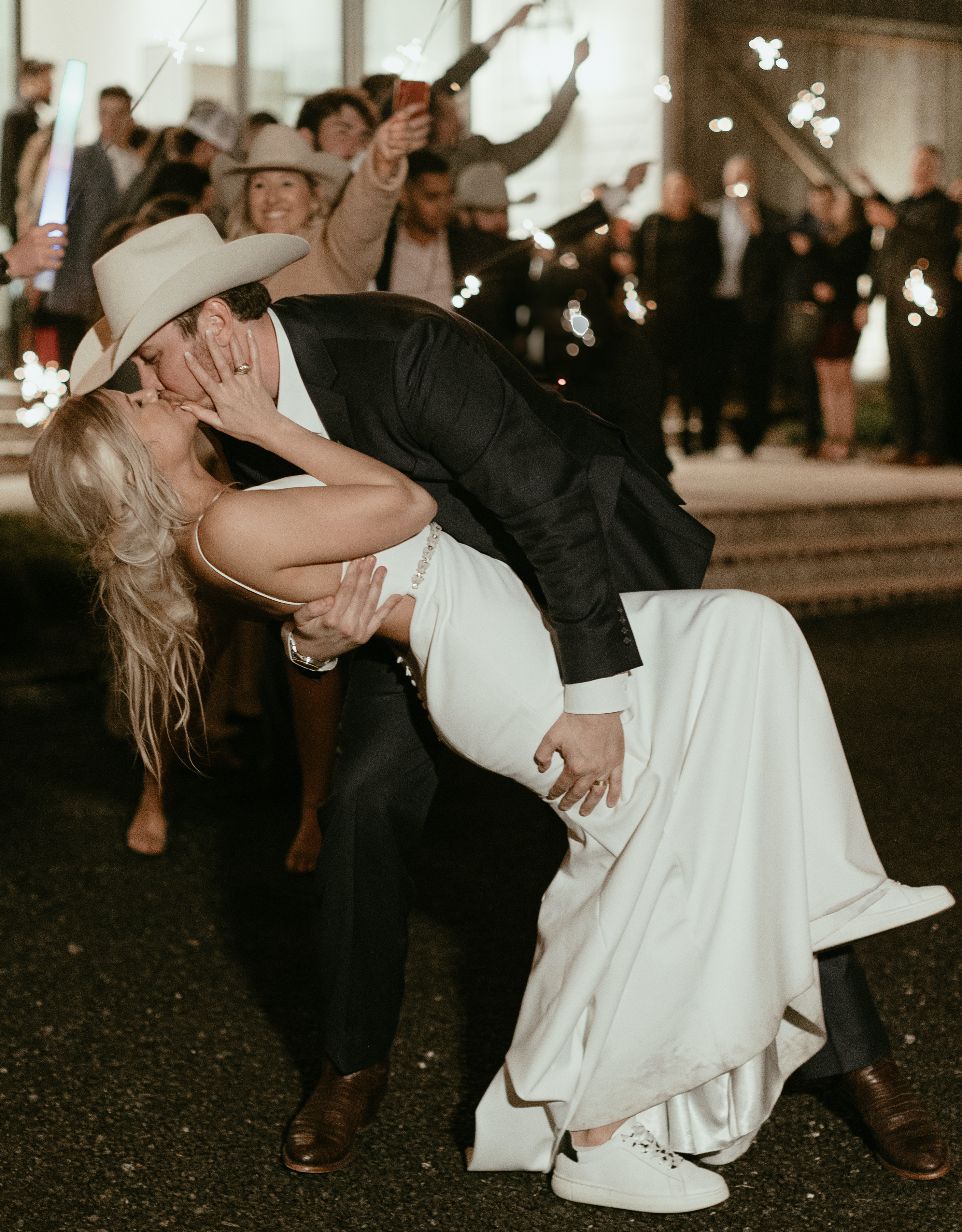 A groom dips his bride outside of their wedding venue while guests hold sparklers in the air. Peach-Toned Countryside wedding.