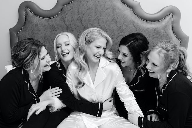 A bride and her bridesmaids sit on a bed smiling wearing silk matching pajamas with their hair and makeup done. 