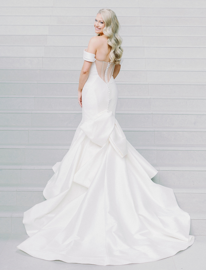 A bride looks over her shoulder and smiles while wearing her white off the shoulder wedding dress. 