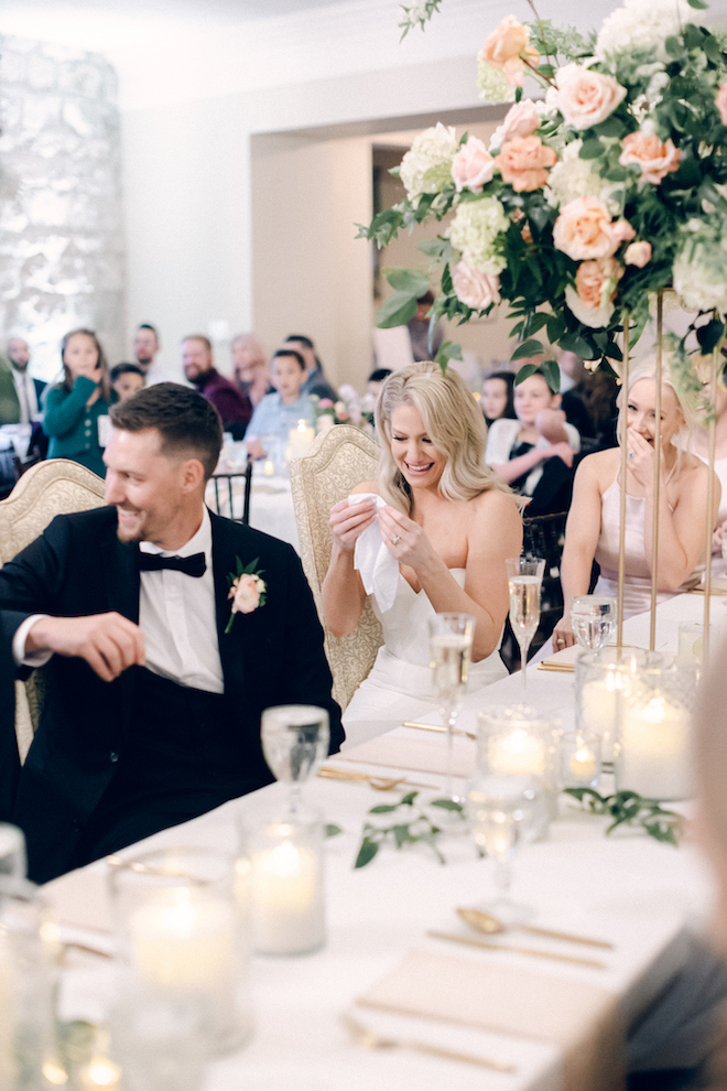 The bride and groom laugh and smile while sitting at a table at their wedding reception. 