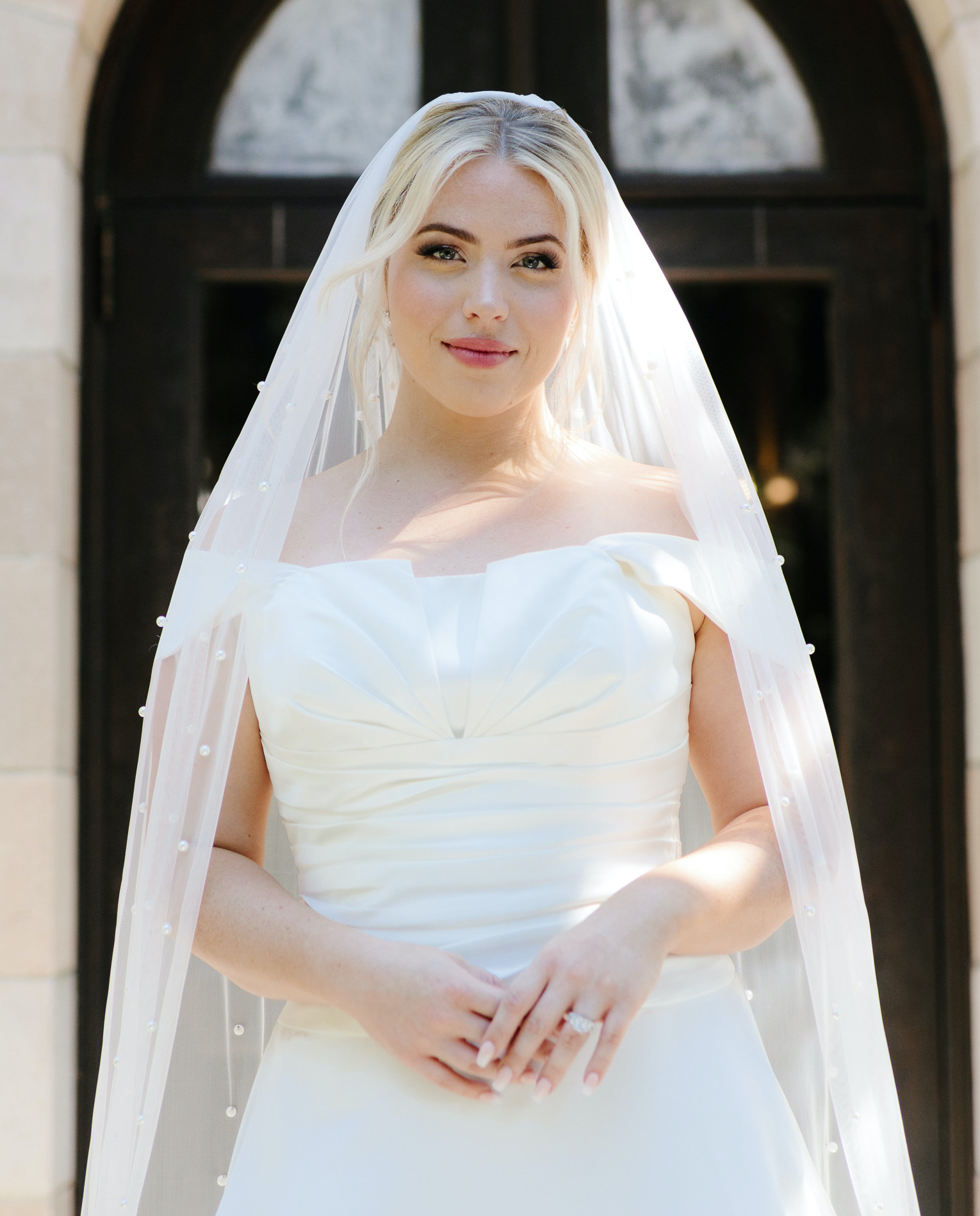 A bride smiles while wearing an off the shoulder white gown and pearl embellished veil in front of a waterfront estate venue in Sarasota, FL