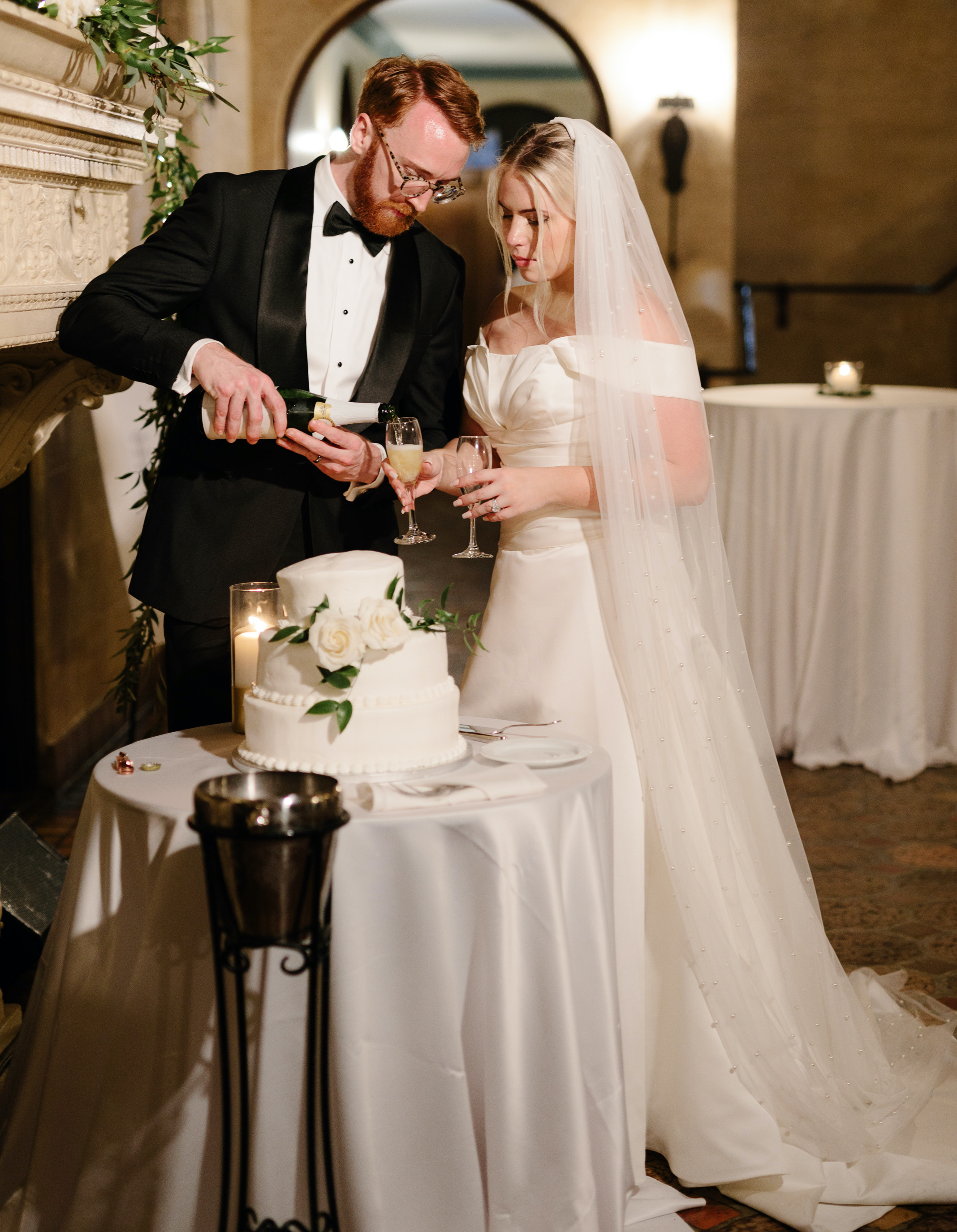 A bride and groom stand by their wedding cake and pour each other champagne.