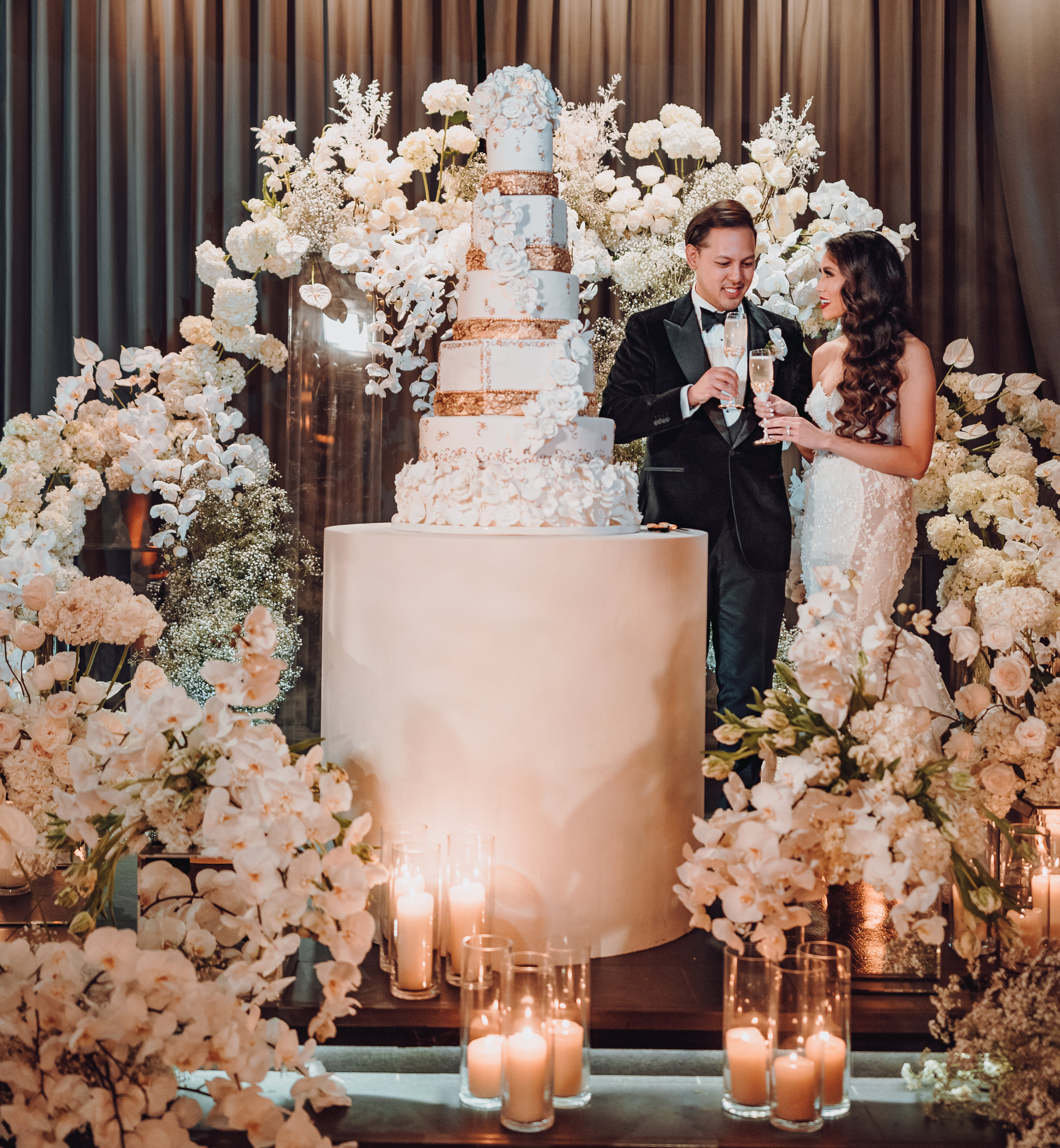 A bride and groom cheers champagne glasses next to their wedding cake. They are surrounded by a floral installation with white flowers.