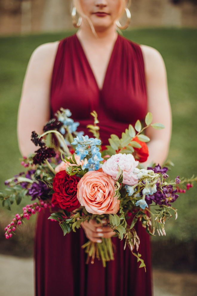 I bridesmaid wearing a burgundy bridesmaid dress holds her bouquet of red, peach, blue, purple and pink flowers. 