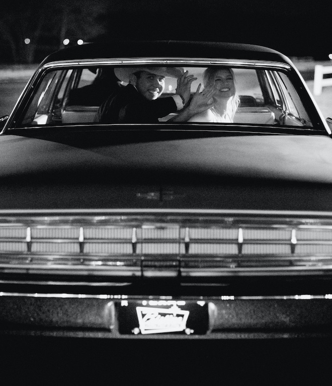 The bride and groom wave goodbye to wedding guests as they exit in a vintage car. 