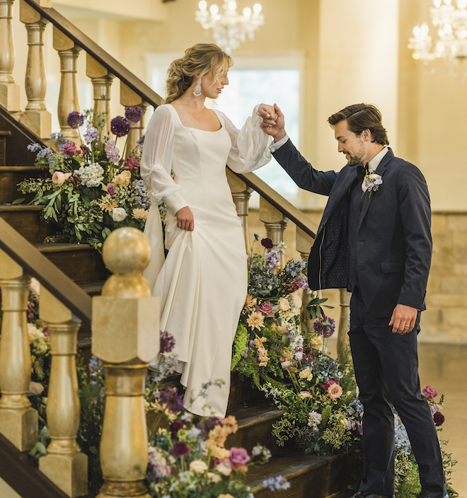 The bride walks down the stairs decorated with pastel flowers and holds the grooms hand. 