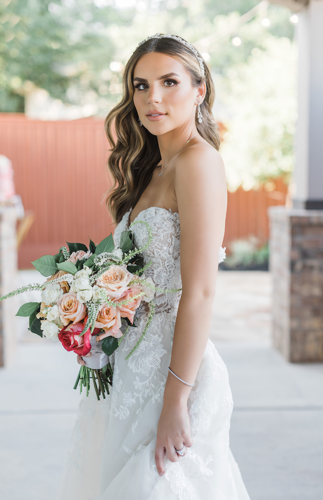 A bride wearing a white, strapless and lace wedding gown holds her pink, peach, white and red wedding bouquet outside the wedding venue, Mara Villa. 