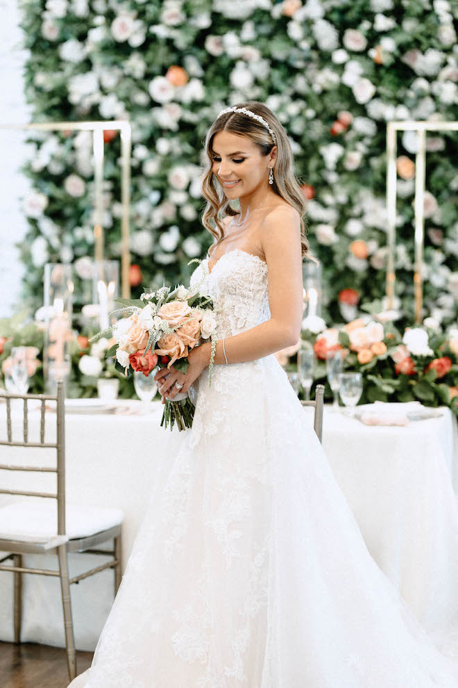 The bride looks down and smiles at her peach, pink and pastel wedding bouquet. 