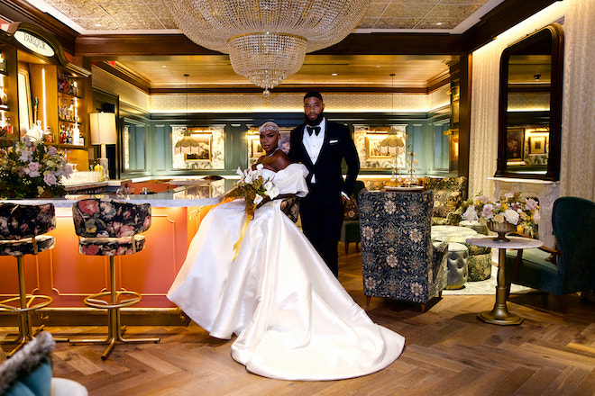 The bride sitting in a chair in the hideaway bar with the groom standing behind her