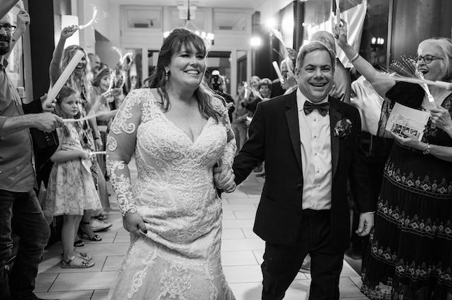 bride and groom hold hands and smile as guests cheer as they exit their wedding venue.