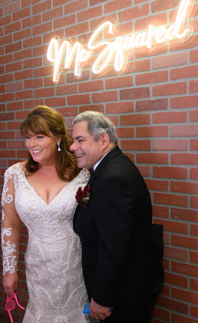 Bride and groom smile aside a neon sign that reads "Mc Squared". 