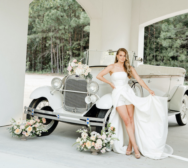 The bride wears a Mikado romper wedding gown infront of a vintage car outside the wedding venue, The Peach Orchard. 